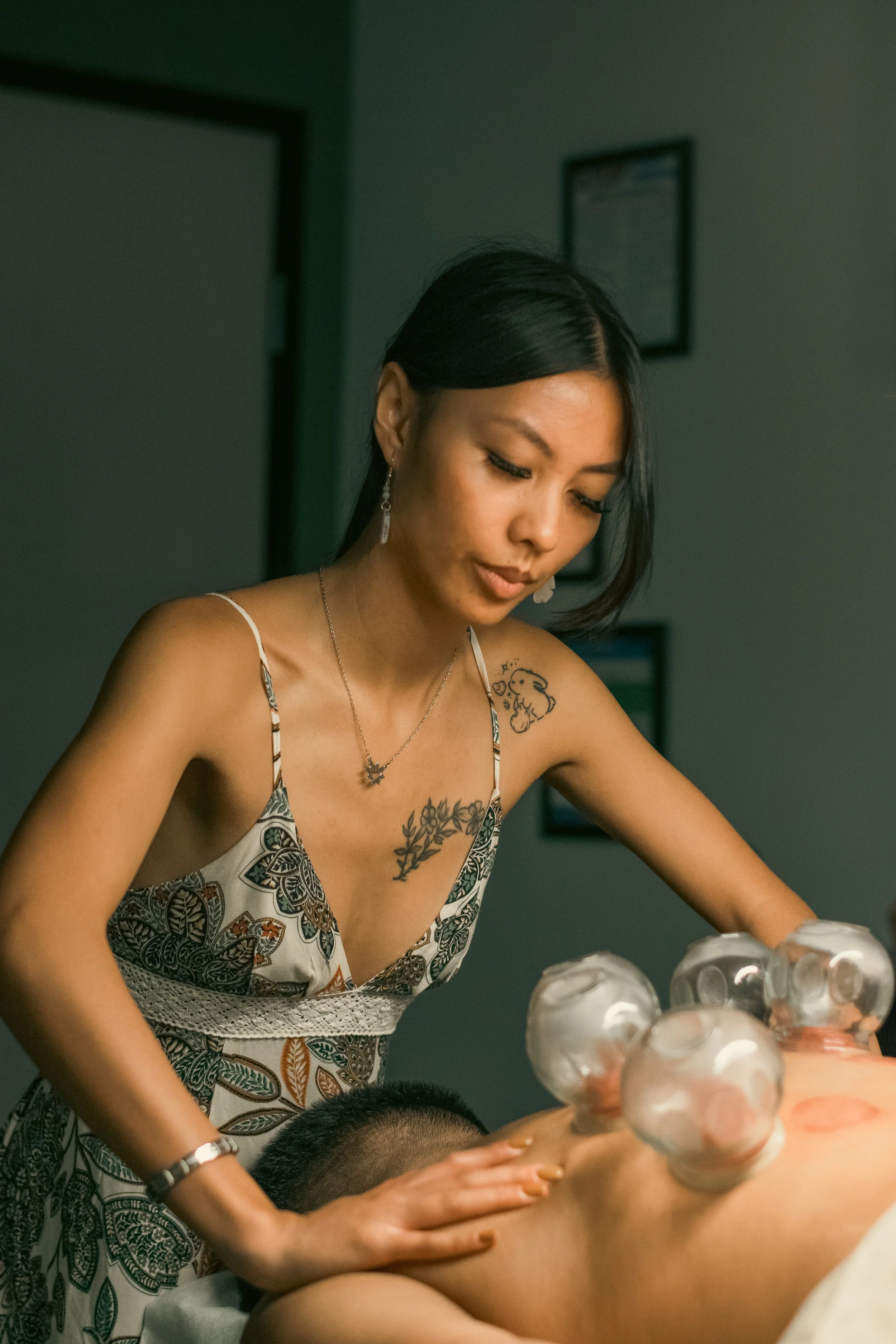 A woman with black hair, tattoos, and wearing jewelry is leaning over a man, massaging his back with ice cubes placed on his back. The scene appears to be in a clinical or therapy setting.