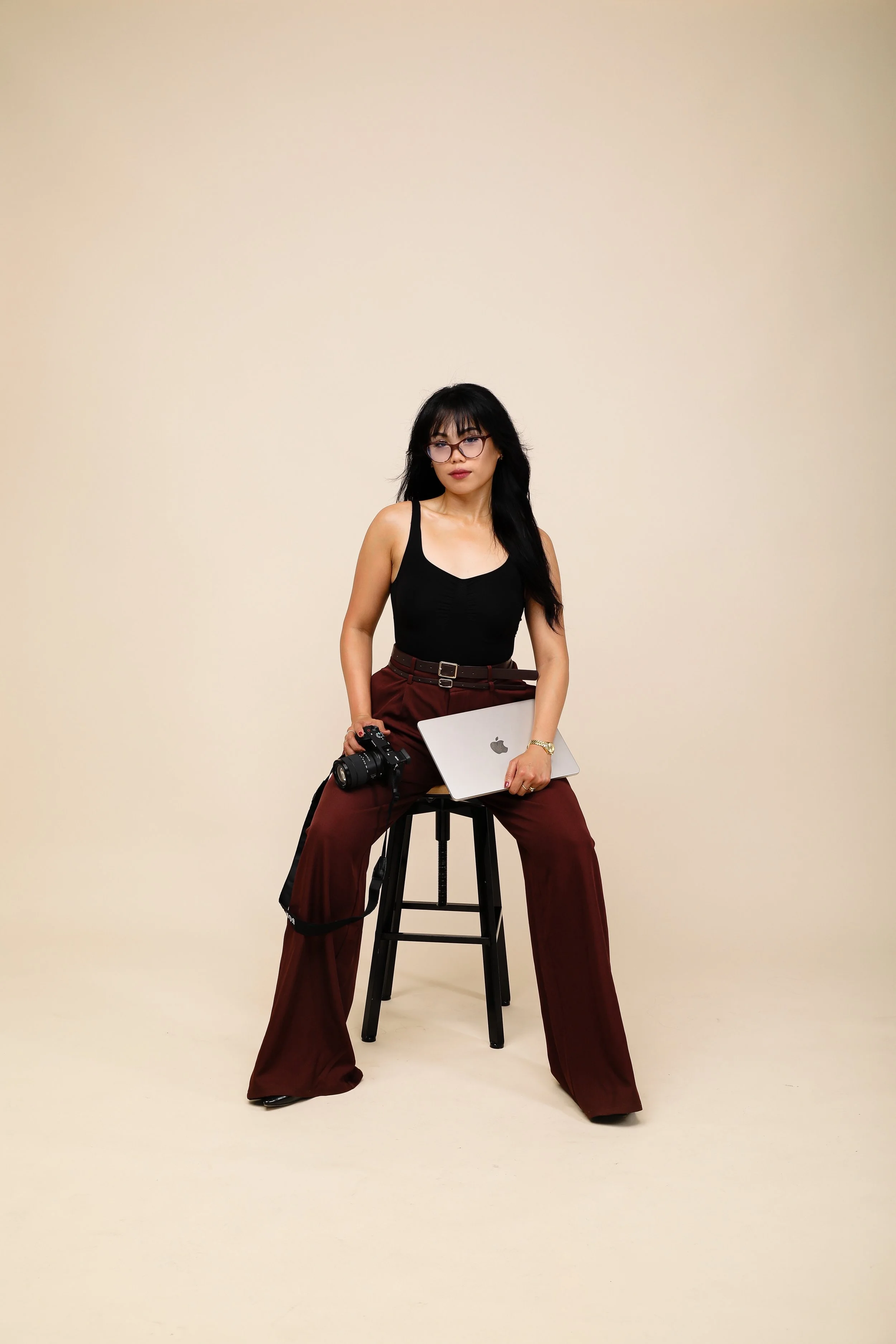 A woman with black hair, glasses, and a black tank top sitting on a tall black stool in front of a plain beige background. She is holding a silver laptop and has a professional camera hanging from her left arm.