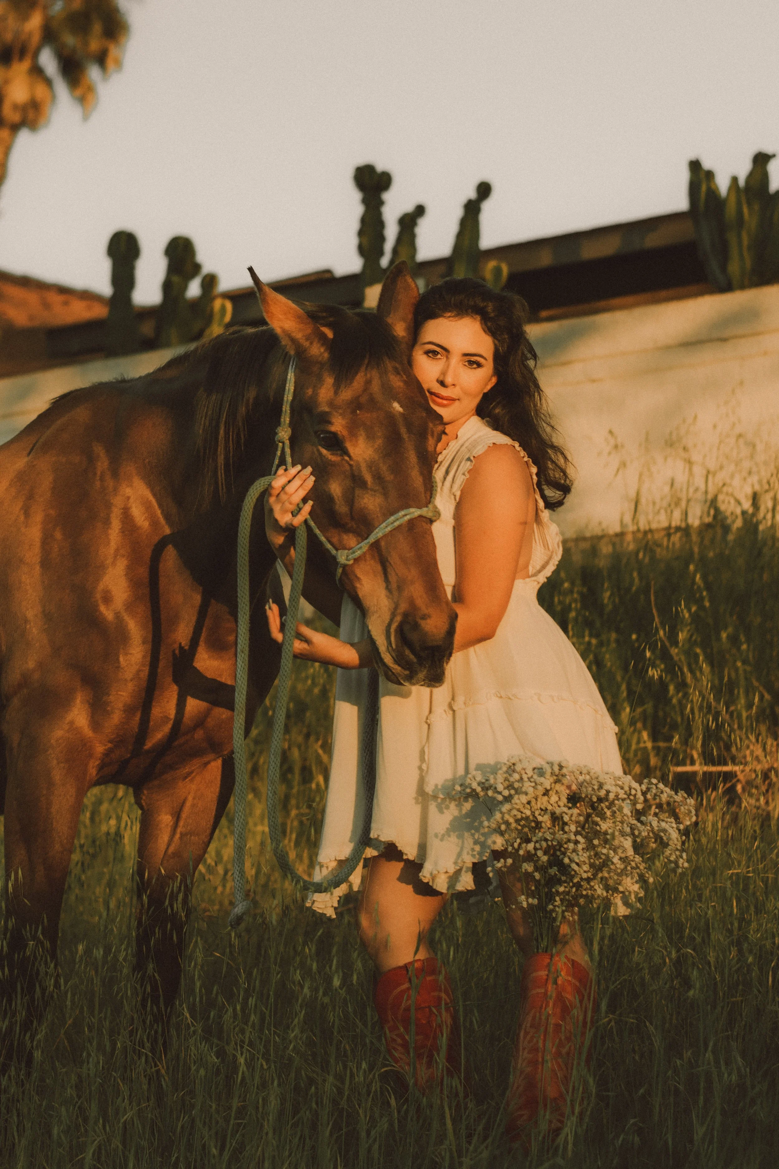 A woman in a white dress standing in a grassy field with a brown horse, holding the horse's reins, during sunset.