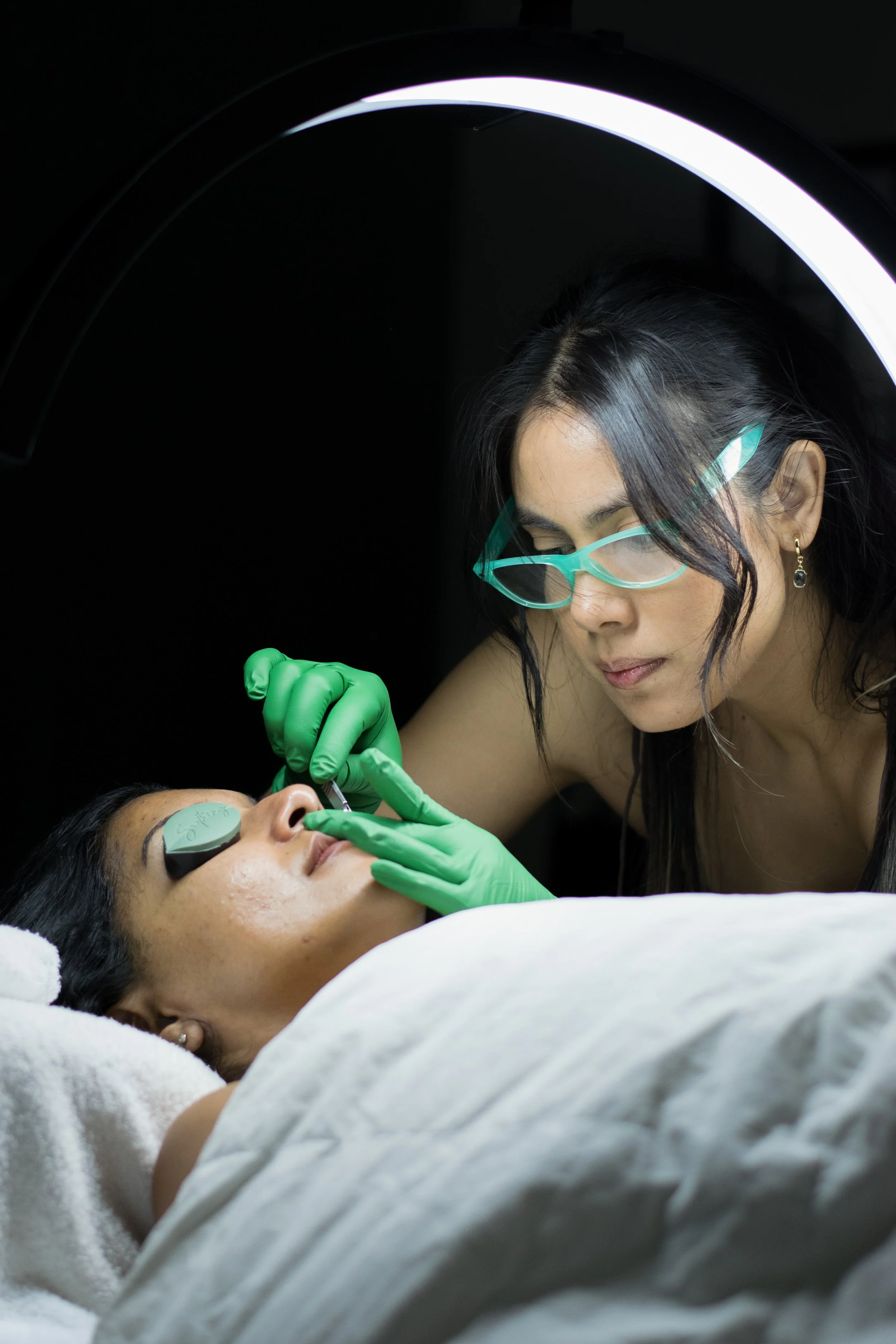 A woman receiving a cosmetic procedure on her nose while lying on a medical bed, with a healthcare professional wearing green gloves, safety glasses, and earrings attending to her in a clinical setting.