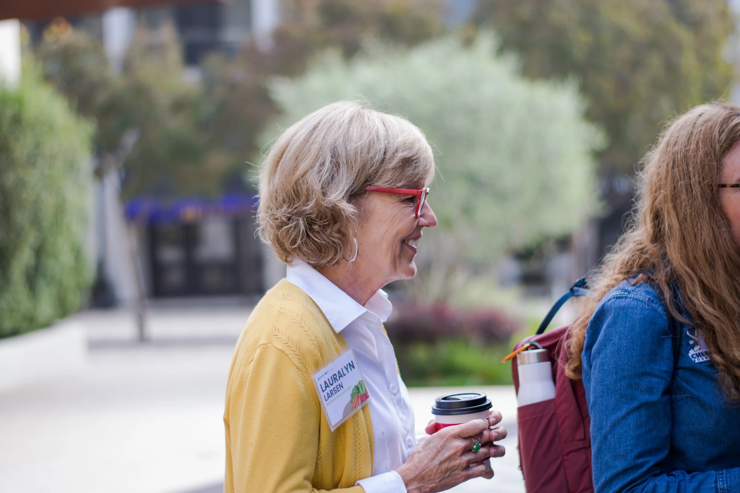 A woman with gray hair and red glasses smiling while holding a coffee cup, standing outdoors with another woman with red hair and a backpack, during daytime at a park or outdoor event.