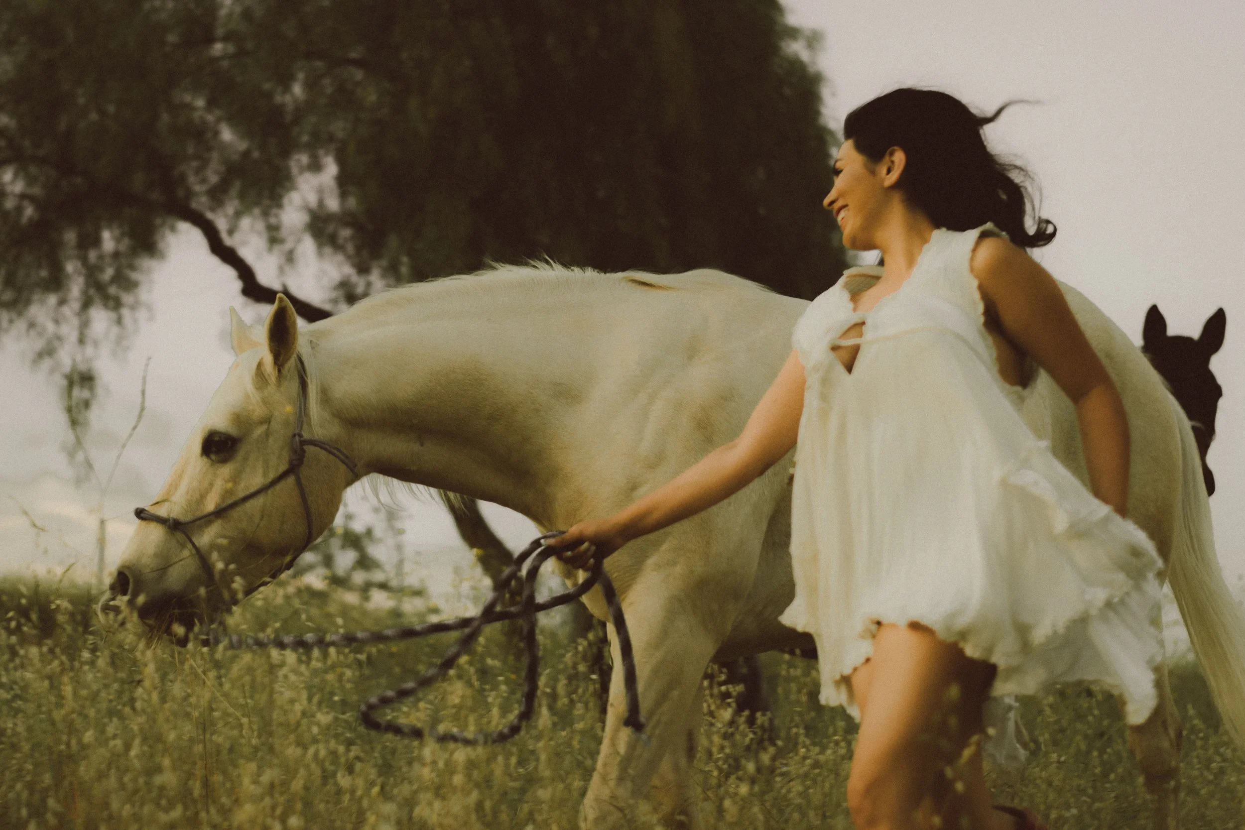A woman in a flowing white dress with cut-out details, standing outdoors and smiling, while holding the reins of a white horse in a grassy field with trees in the background.