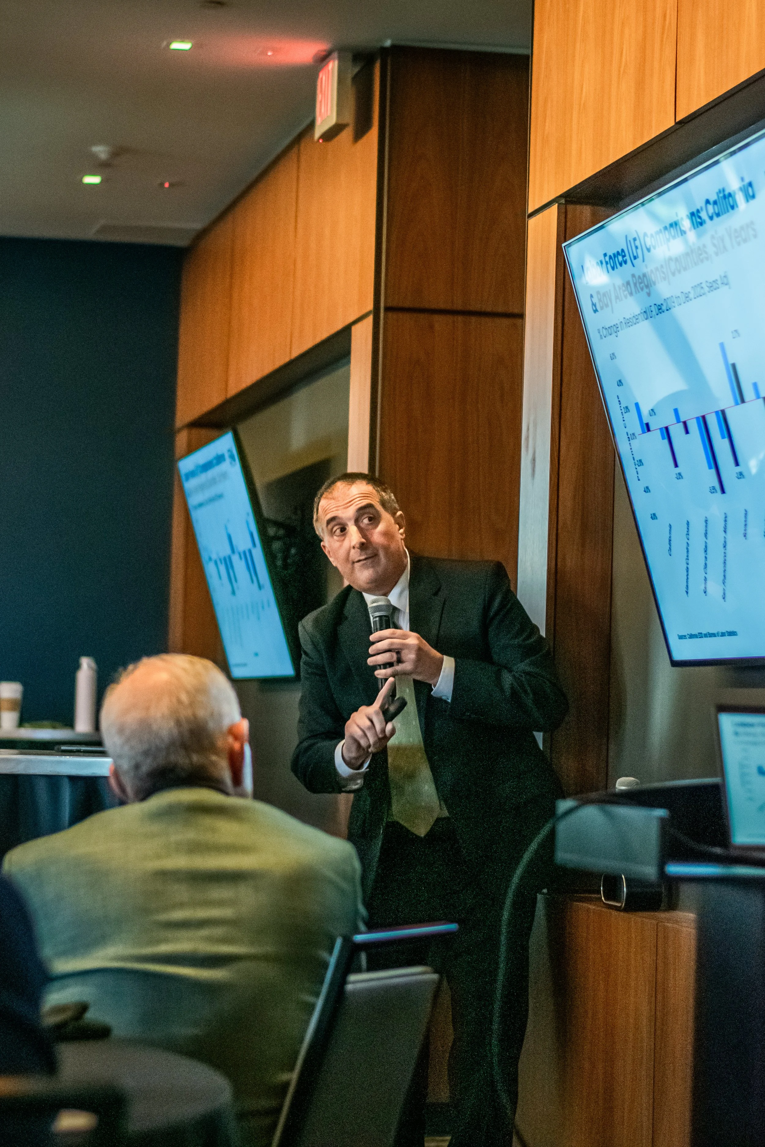 A man in a suit holding a microphone presents in front of an audience in a conference room, with several screens displaying charts and text behind him.