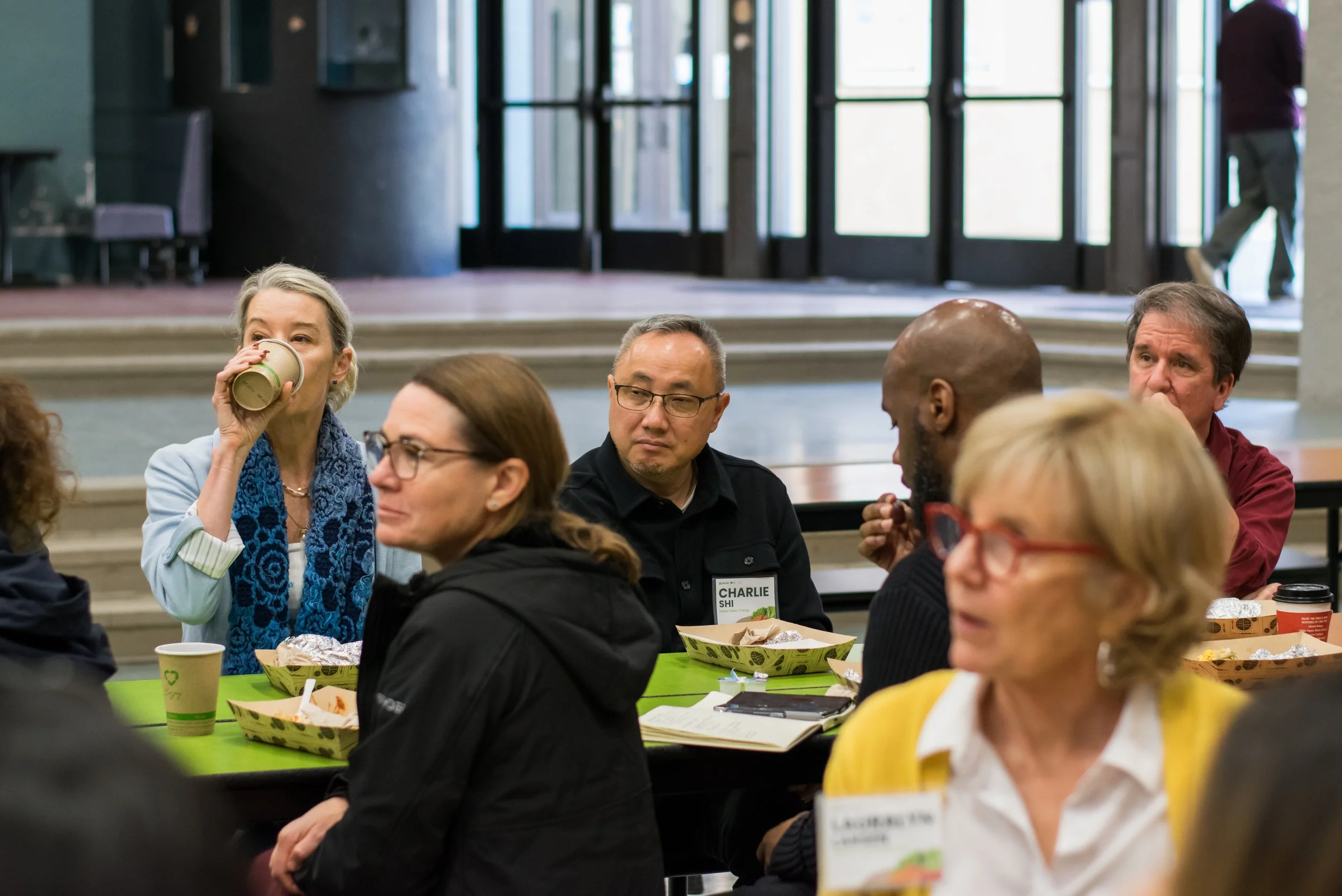 Group of diverse people sitting at a table in a brightly lit indoor space, some eating and drinking, engaged in conversation during a social or professional gathering.