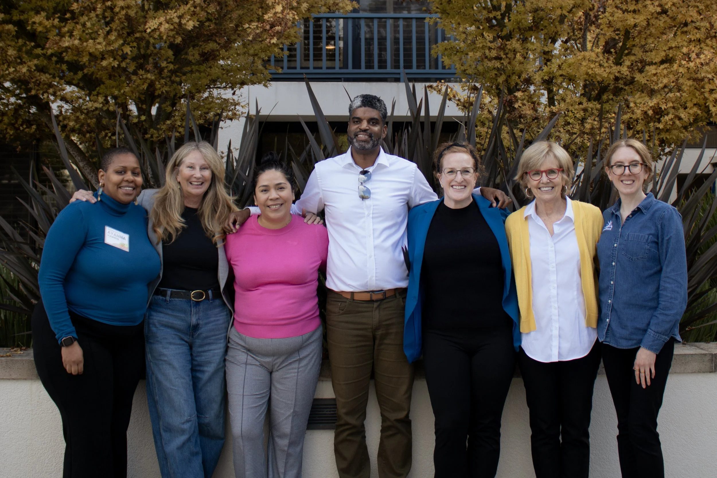Group of seven diverse people standing outdoors in front of autumn trees, smiling, with arms around each other's shoulders.