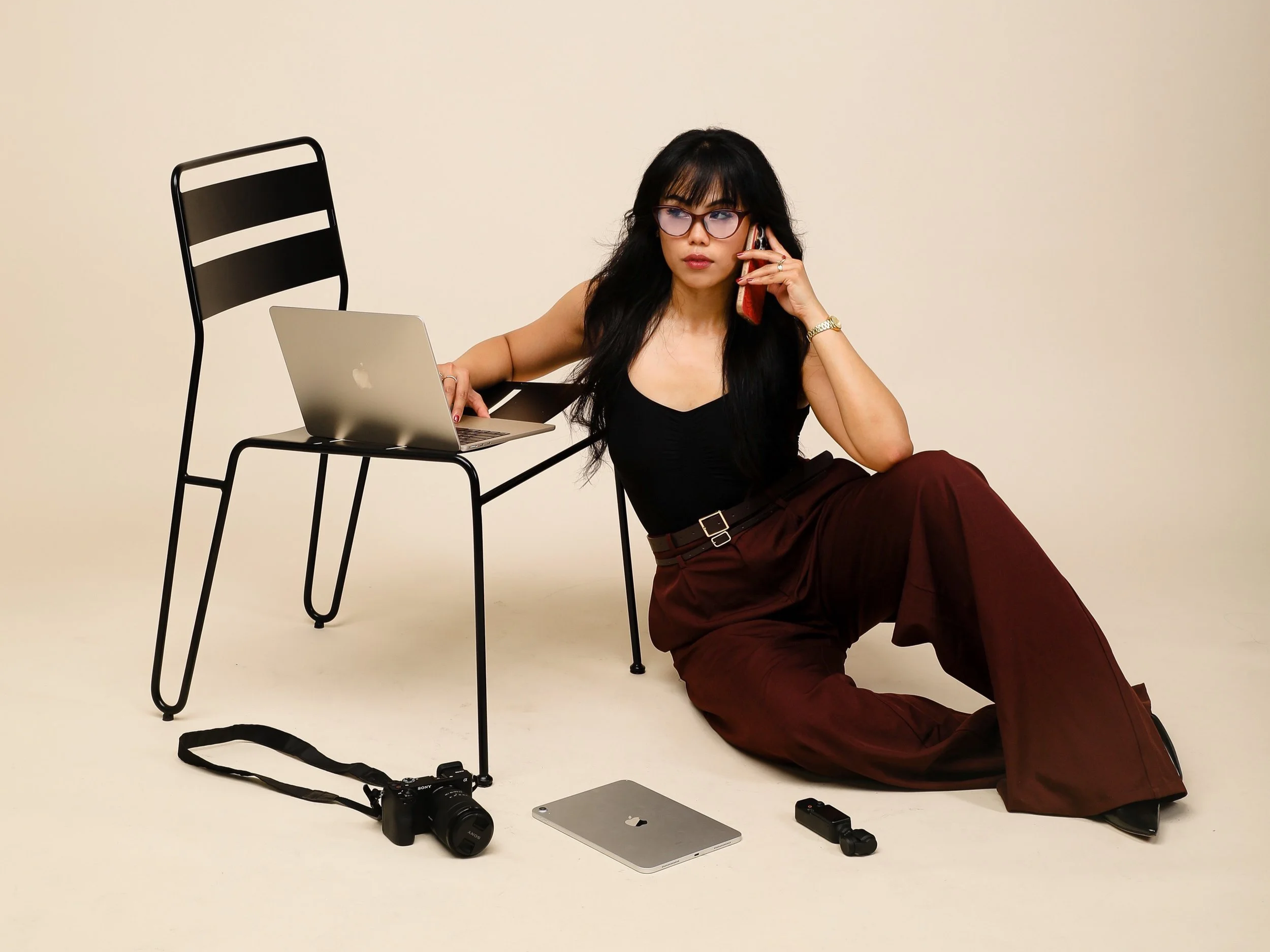 Woman with black hair and glasses sitting on the floor, talking on a cellphone, with photography equipment and electronic devices around her, including camera, laptop, and camera accessories, in a studio with a plain beige background.