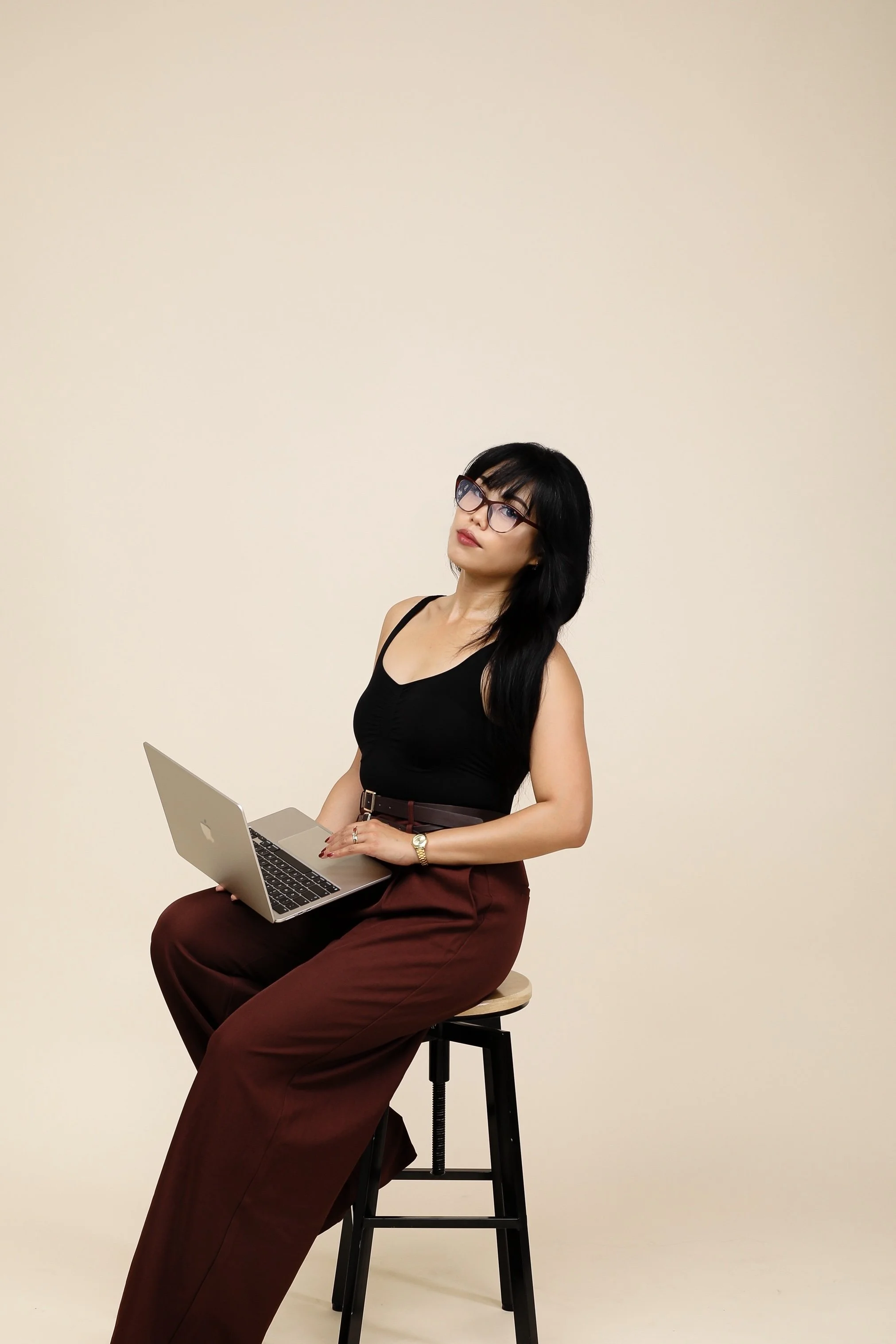 A woman with black hair, glasses, wearing a black top and maroon pants, sitting on a stool holding a laptop, against a plain beige background.