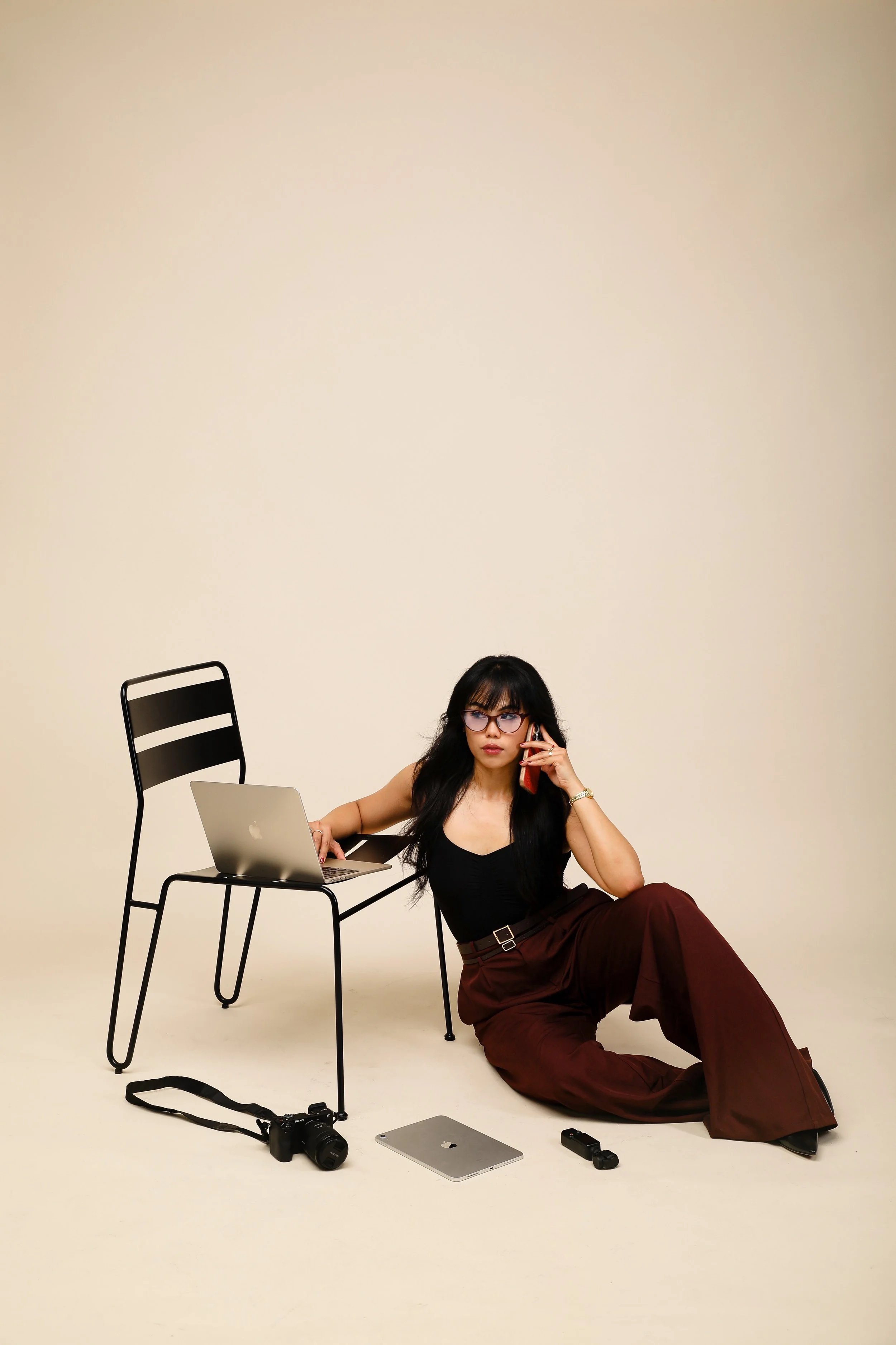 A woman with glasses and long black hair, sitting on the floor with her back supported by a chair, holding a phone to her ear, and surrounded by various electronic devices, including a laptop, tablet, camera, and camera accessories, against a plain beige background.