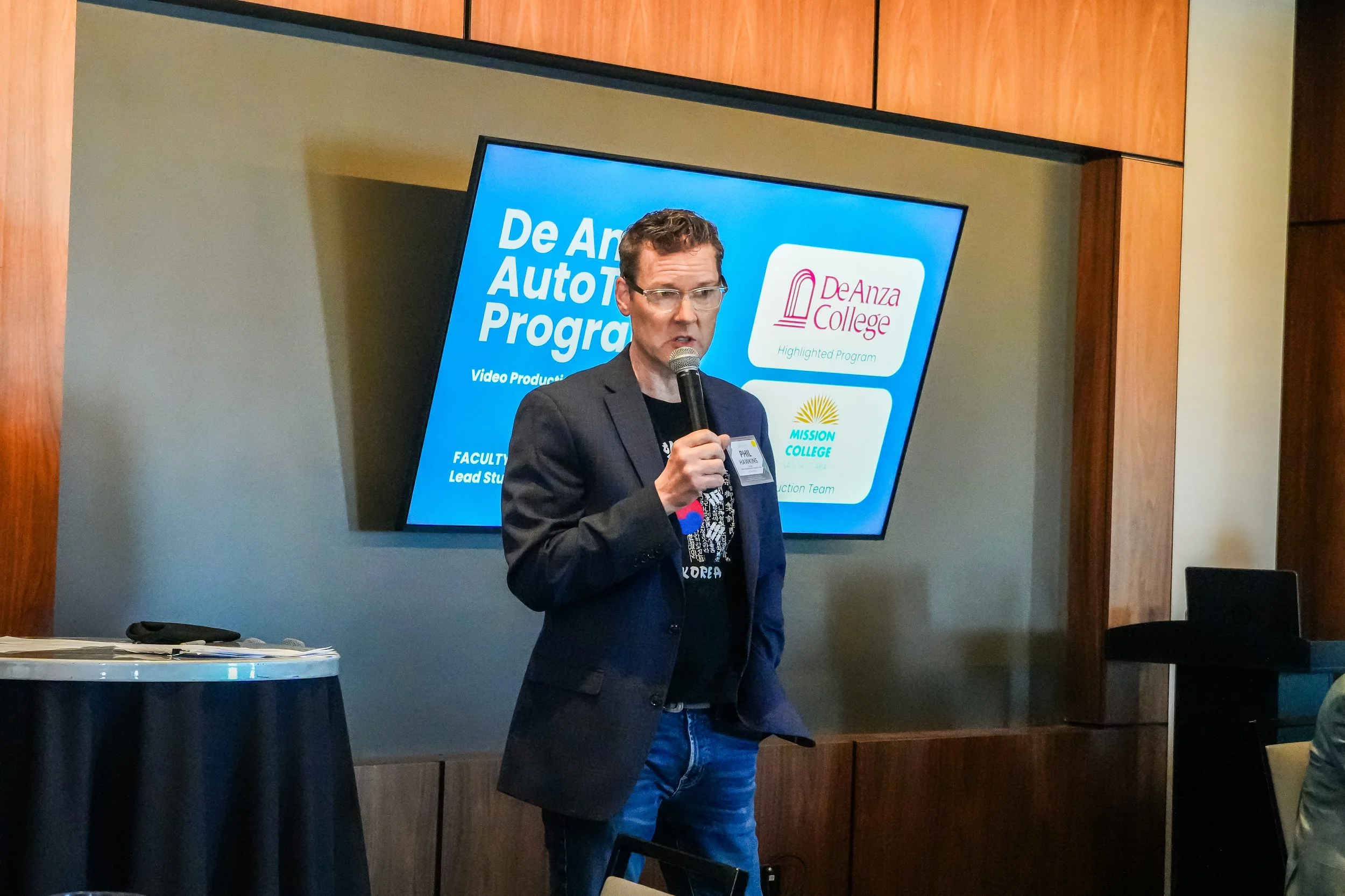 A man is speaking into a microphone at a presentation or event. He is wearing glasses, a blazer, and a black T-shirt. Behind him is a large screen displaying the text 'De Anza Autobody Program' and logos for De Anza College and Mission College.