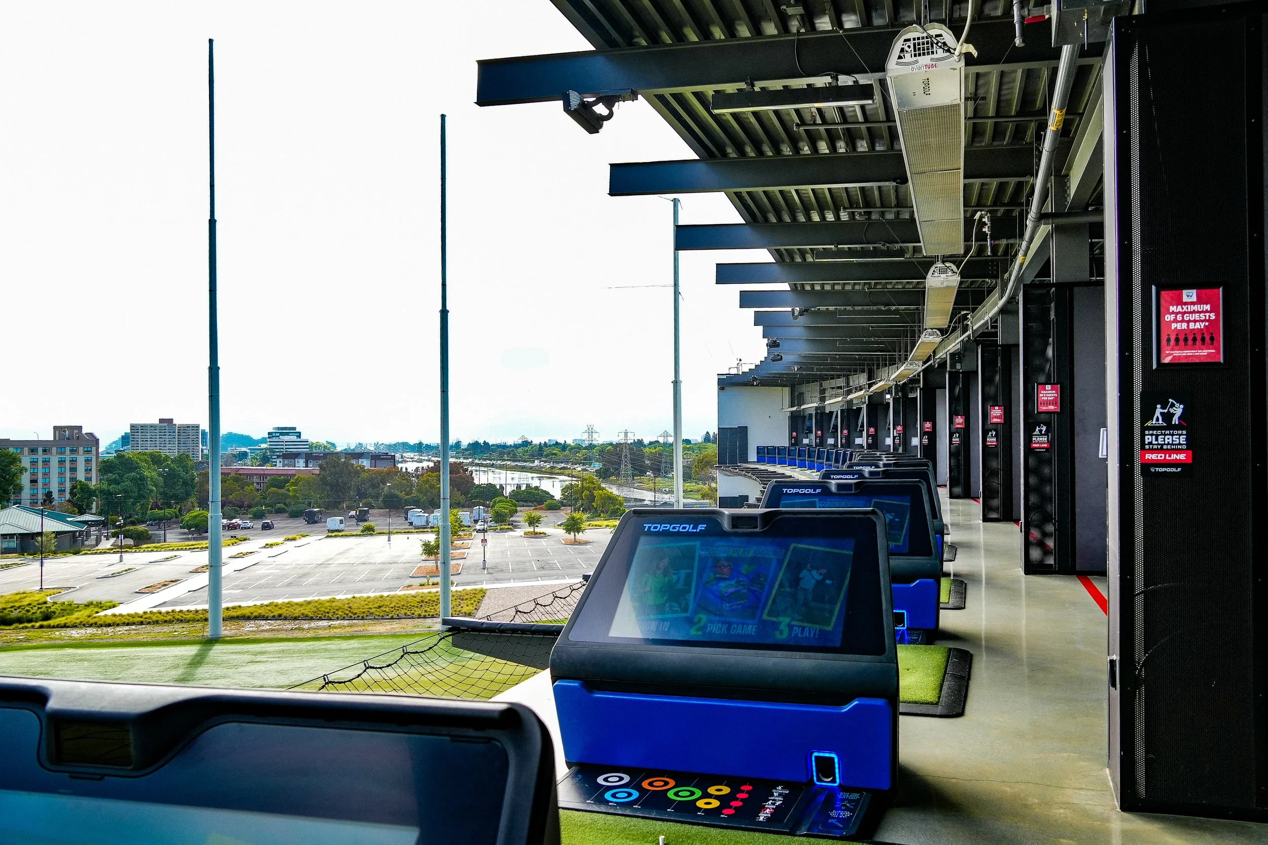 Inside view of a driving range with electronic golf game screens and a view of the outside parking lot and cityscape with trees and buildings in the background.