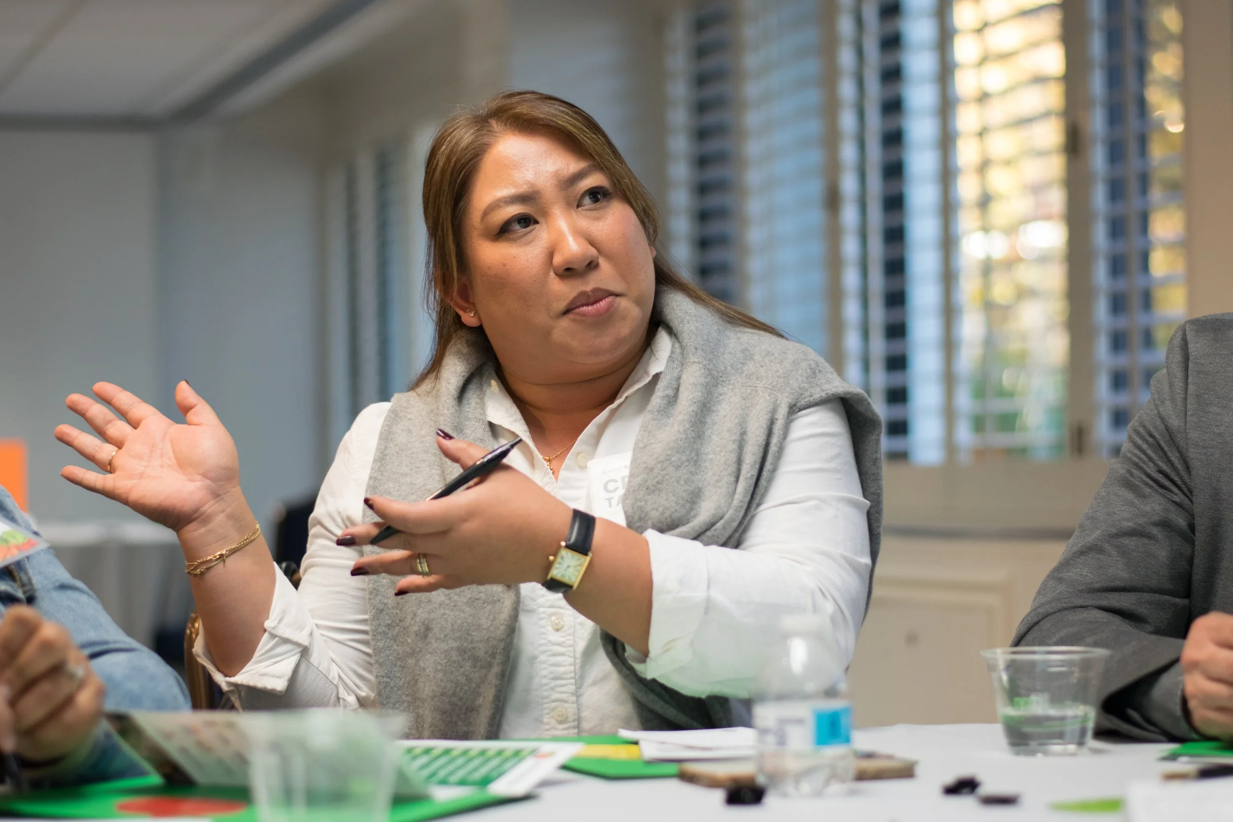 A woman in a white shirt and gray vest is engaged in a discussion at a meeting, gesturing with her hands. She has brown hair and is wearing a gold bracelet and a watch. Other people are partially visible around the table, with a background of windows