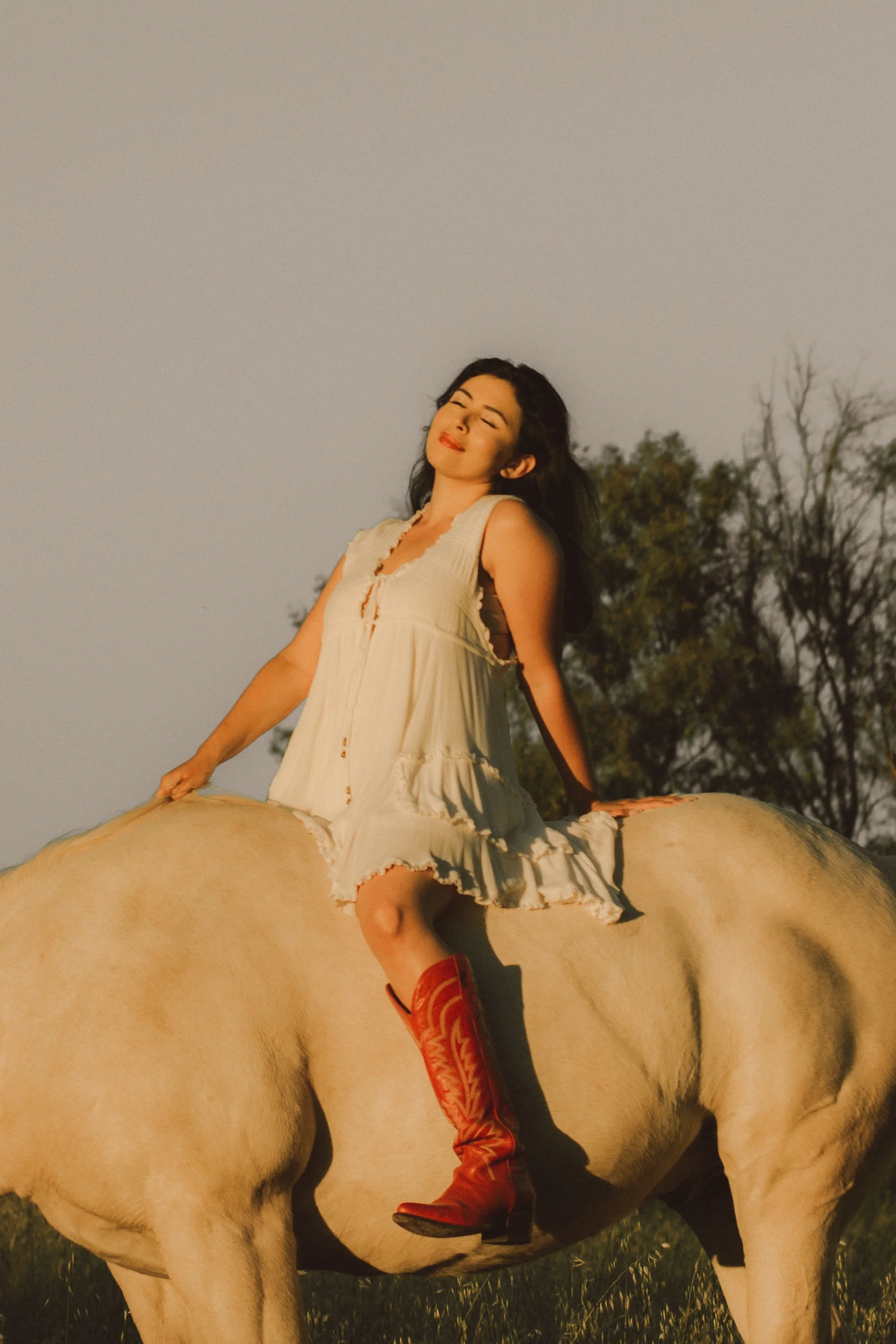 Woman with long black hair, wearing red boots and a cream dress, sitting on a white horse in an outdoor setting with trees in the background and a clear sky.
