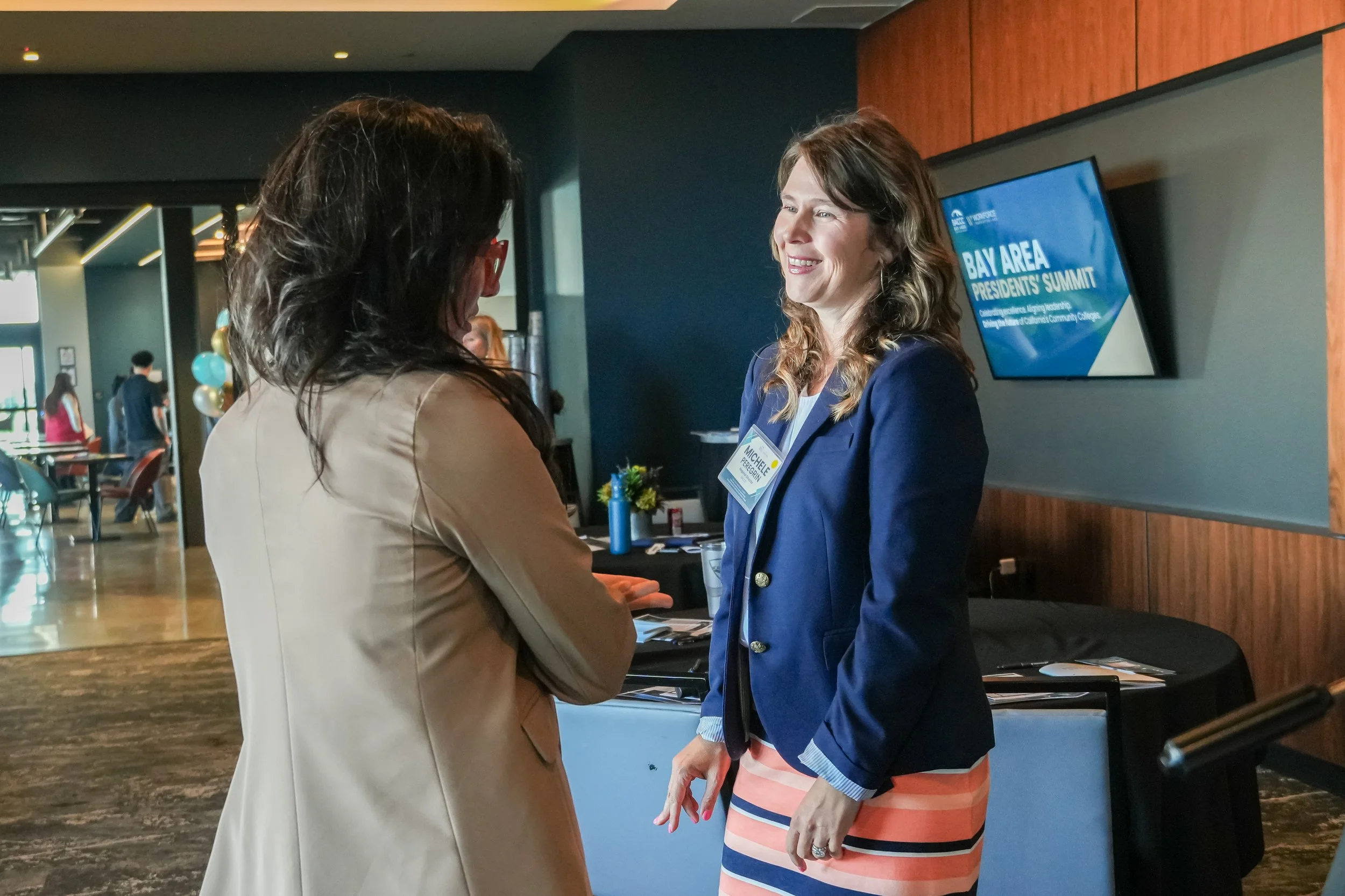 Two women engaged in conversation at a professional event inside a conference center, with a digital display in the background reading 'Bay Area Presidents' Summit.'