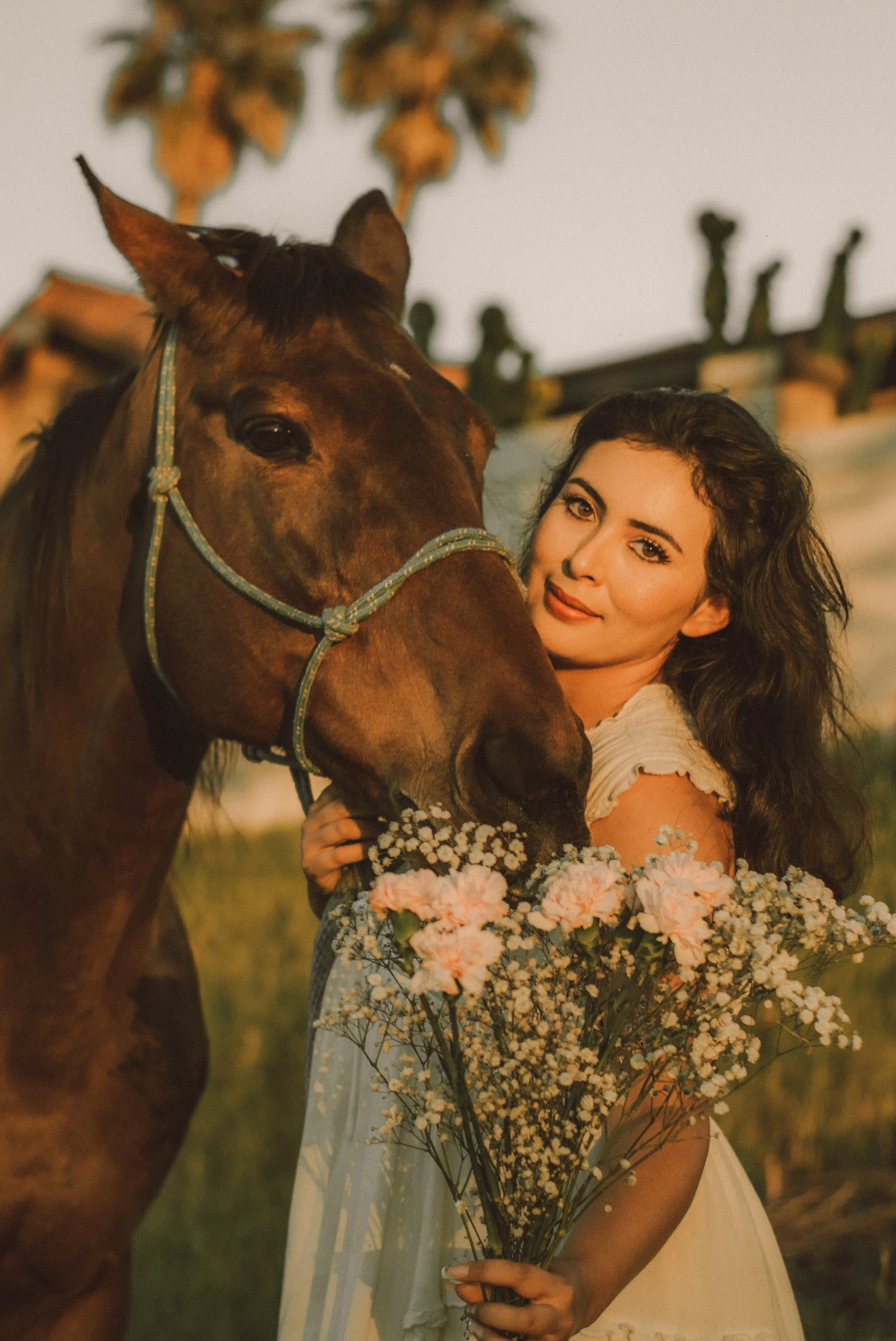 A woman with long dark hair holding a large bouquet of pink and white flowers, standing next to a brown horse with a blue halter, in a sunny outdoor setting with a building and plants in the background.
