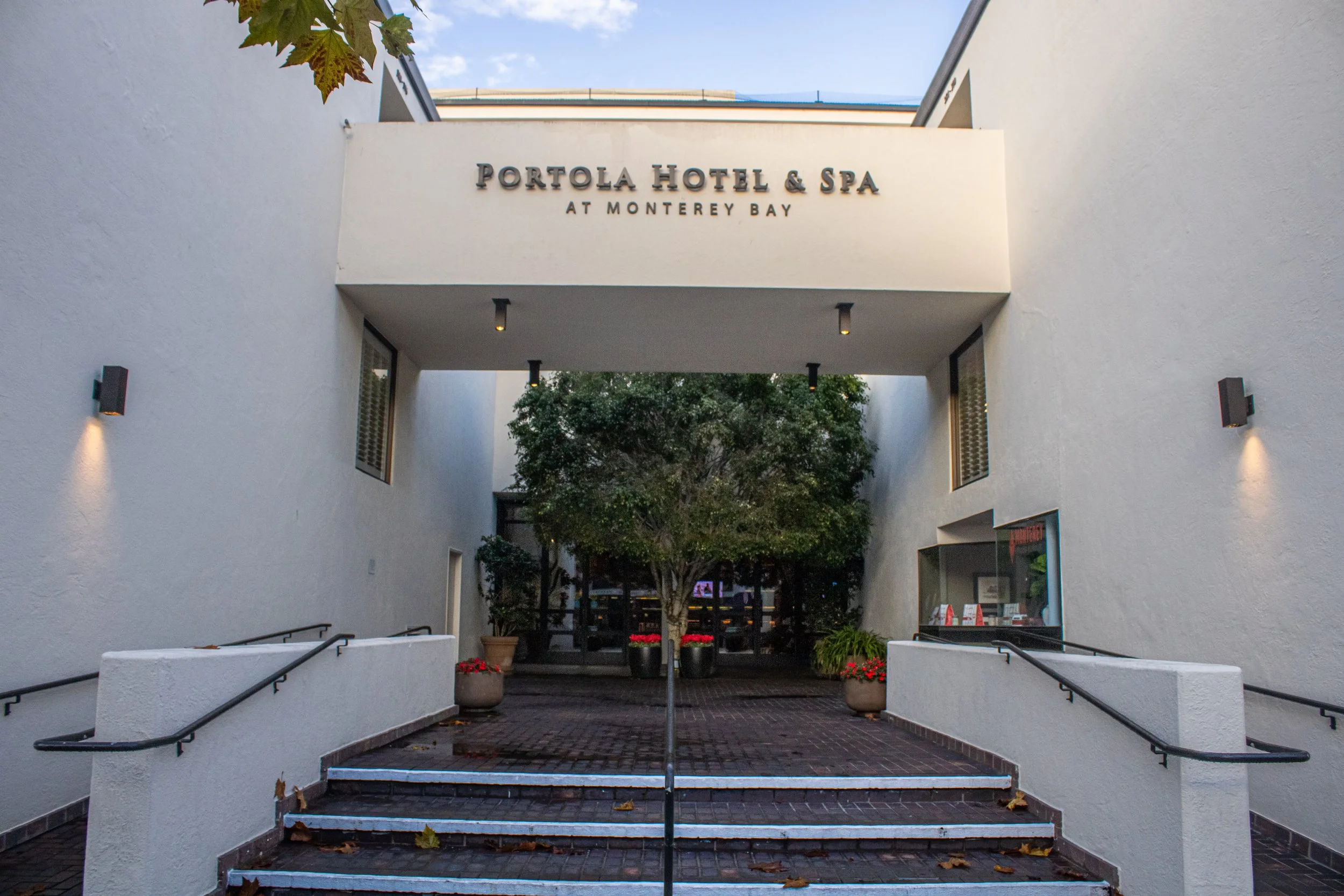 Entrance to Portola Hotel & Spa at Monterey Bay with white walls, illuminated wall lights, stairs, potted plants, and trees outside, under a partly cloudy sky.