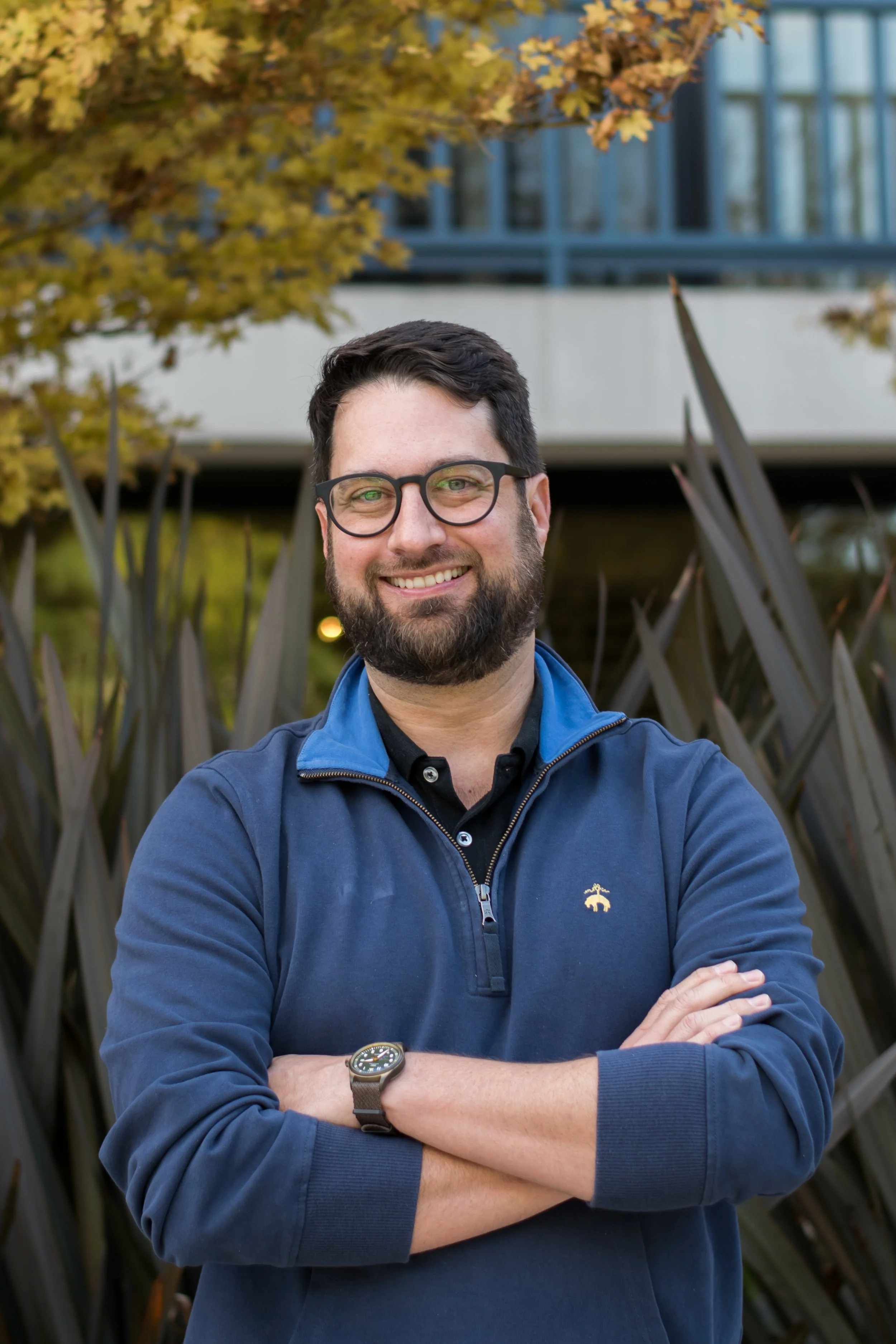 A smiling man with glasses and a beard, wearing a blue jacket and watch, standing outdoors in front of an outdoor plant and a building with blue-framed windows.