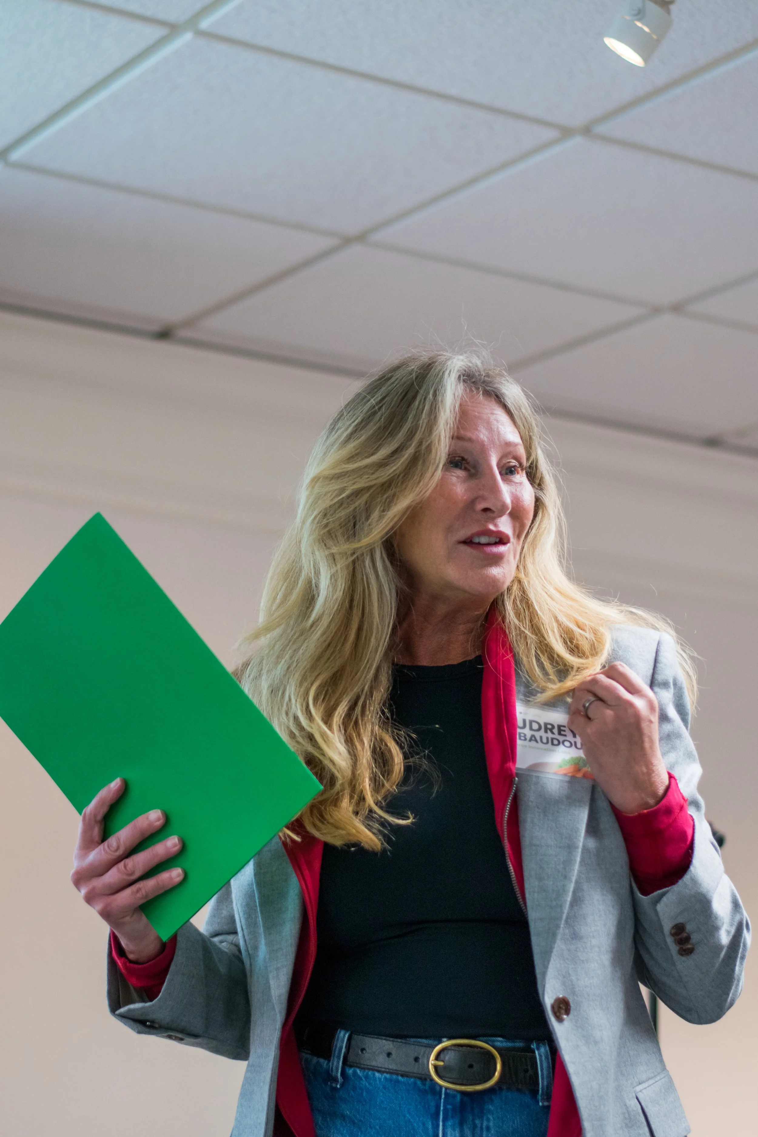 A woman with long blonde hair wearing a gray blazer, black shirt, and jeans holding a green folder in one hand and a small object in her other hand, standing in an indoor setting with a drop ceiling and a spotlight above.