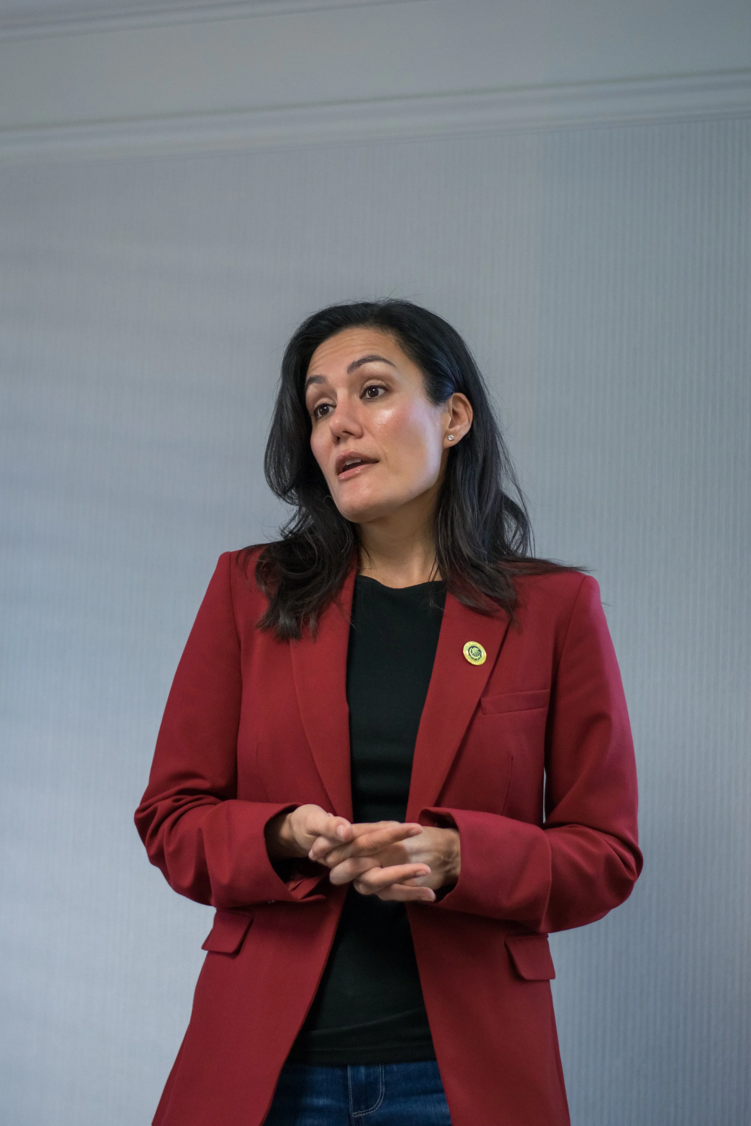 A woman with dark hair wearing a red blazer, black shirt, and jeans, standing in front of a plain light-colored wall, speaking or making a gesture.