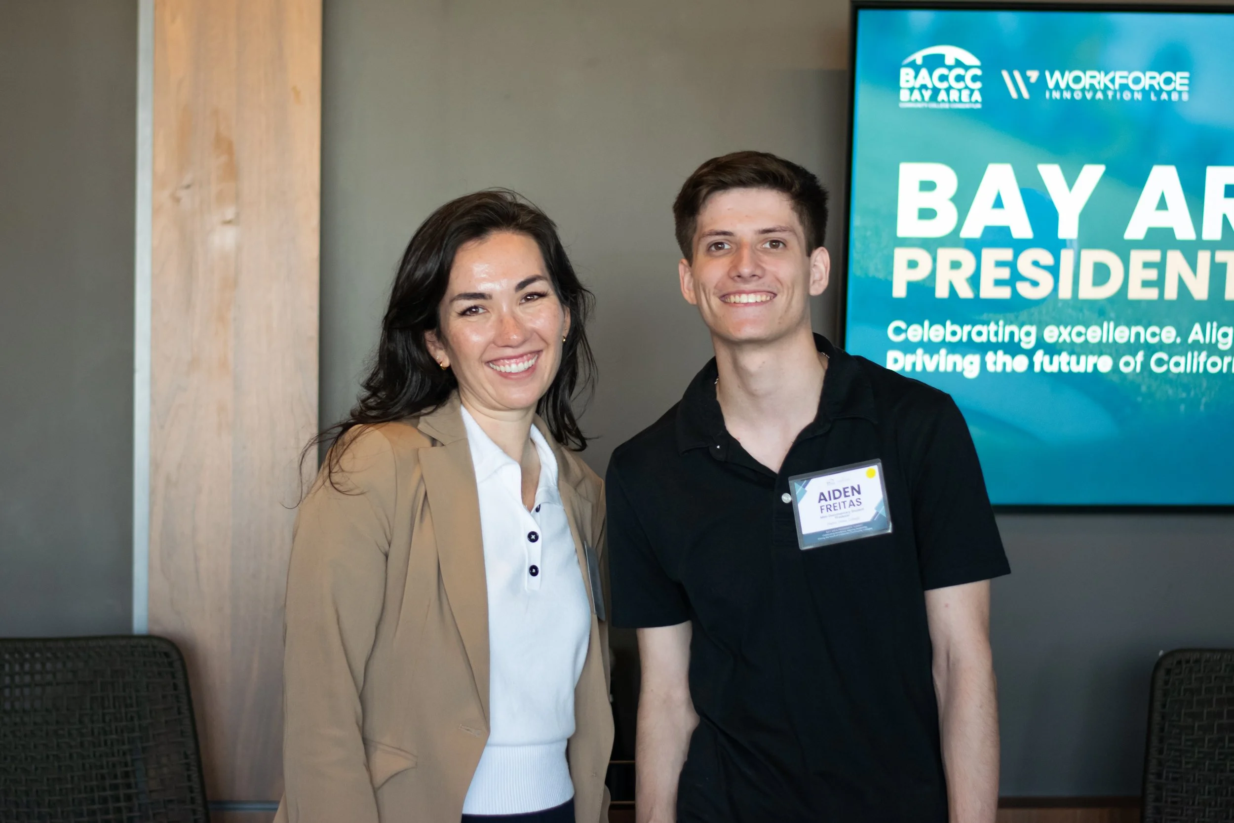 A woman and a young man standing together at an indoor event, smiling at the camera. The woman has dark hair, is wearing a beige blazer and white shirt. The man, identified by a name tag as Aiden Freitas, has short dark hair, is wearing a black shirt
