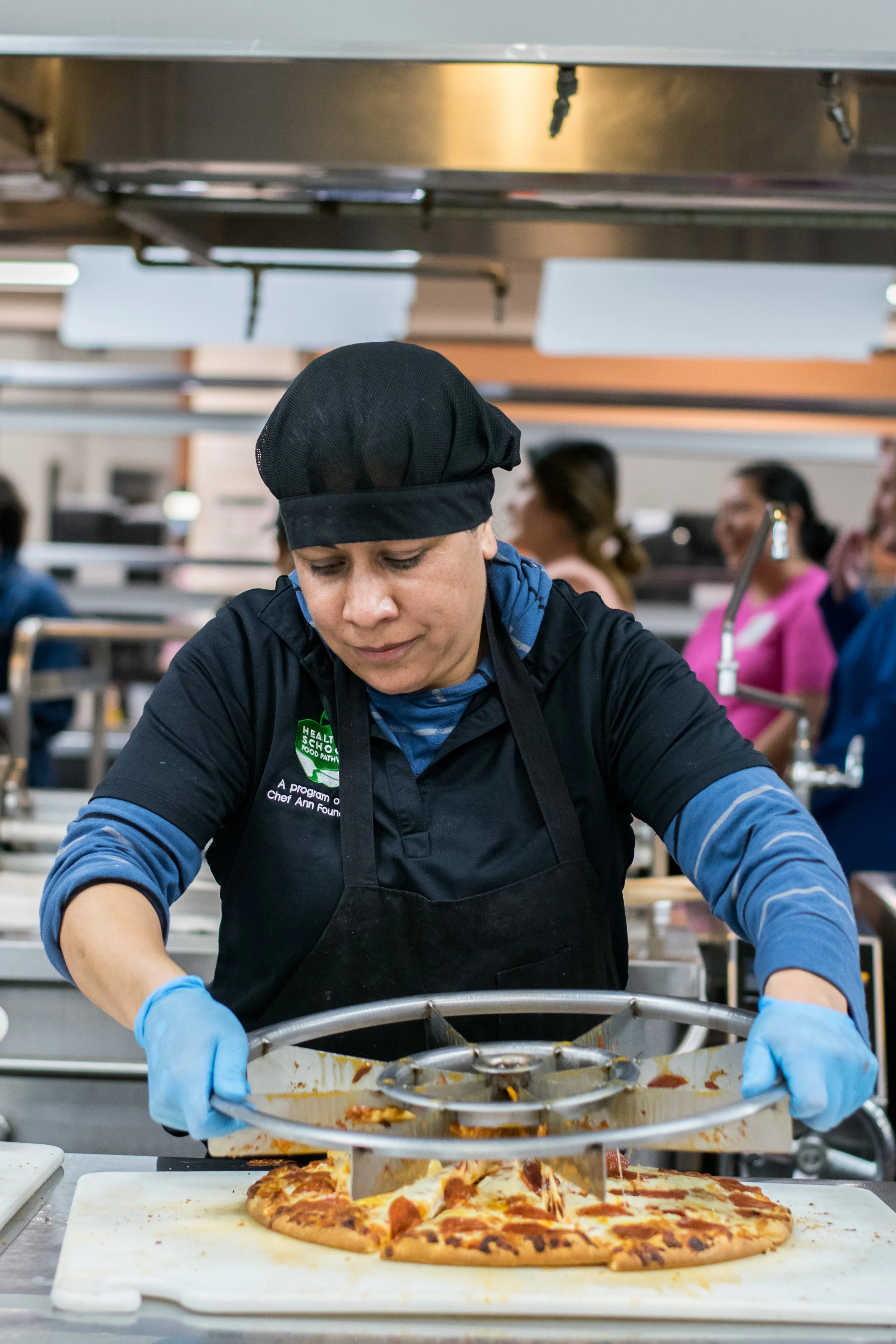 A woman in a black chef hat and blue gloves preparing pizza in a commercial kitchen.