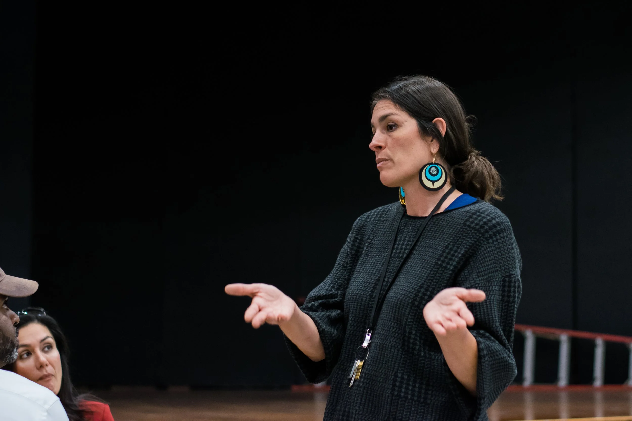 A woman with dark hair wearing large colorful earrings is engaged in conversation with people sitting in front of her at an indoor event.