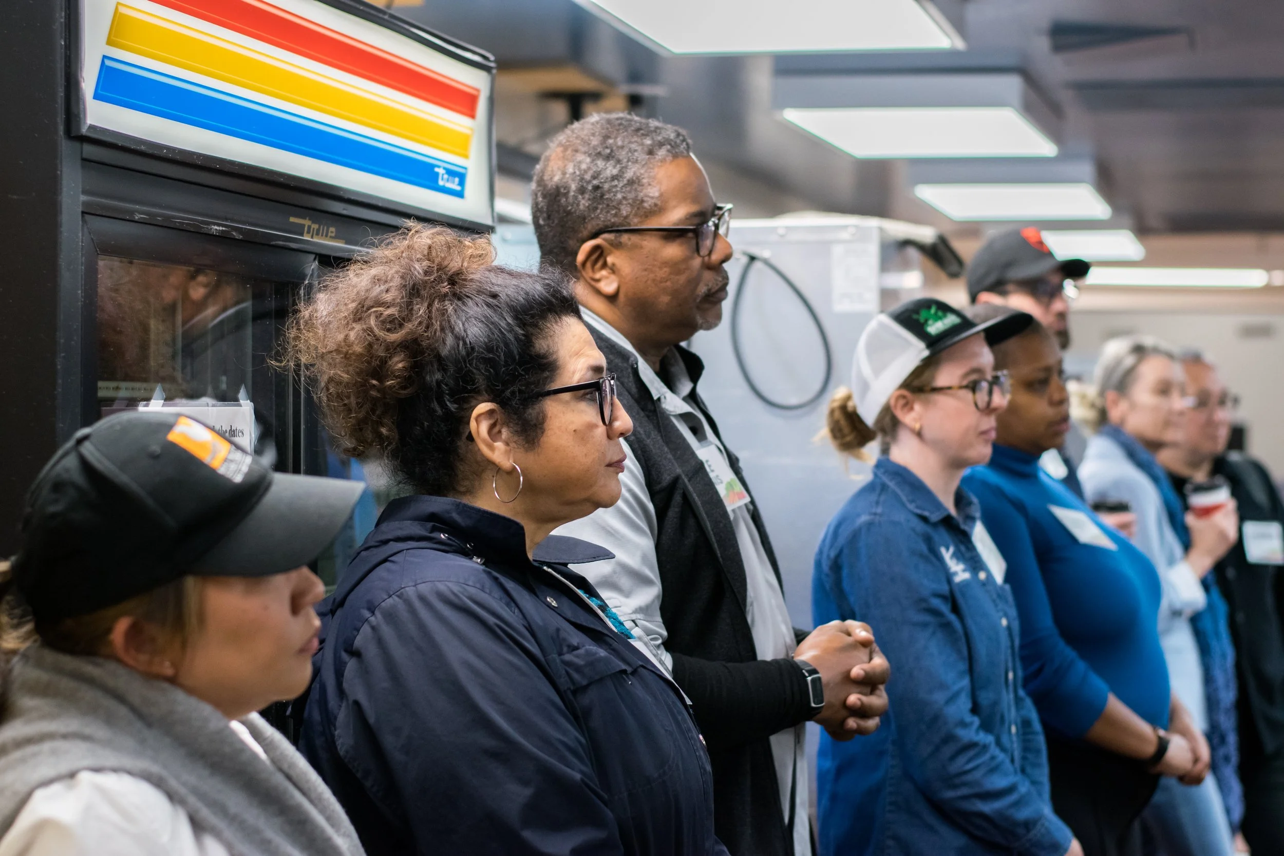 A diverse group of people standing in a line, with eyes closed and hands clasped, possibly praying or meditating, in an indoor setting.