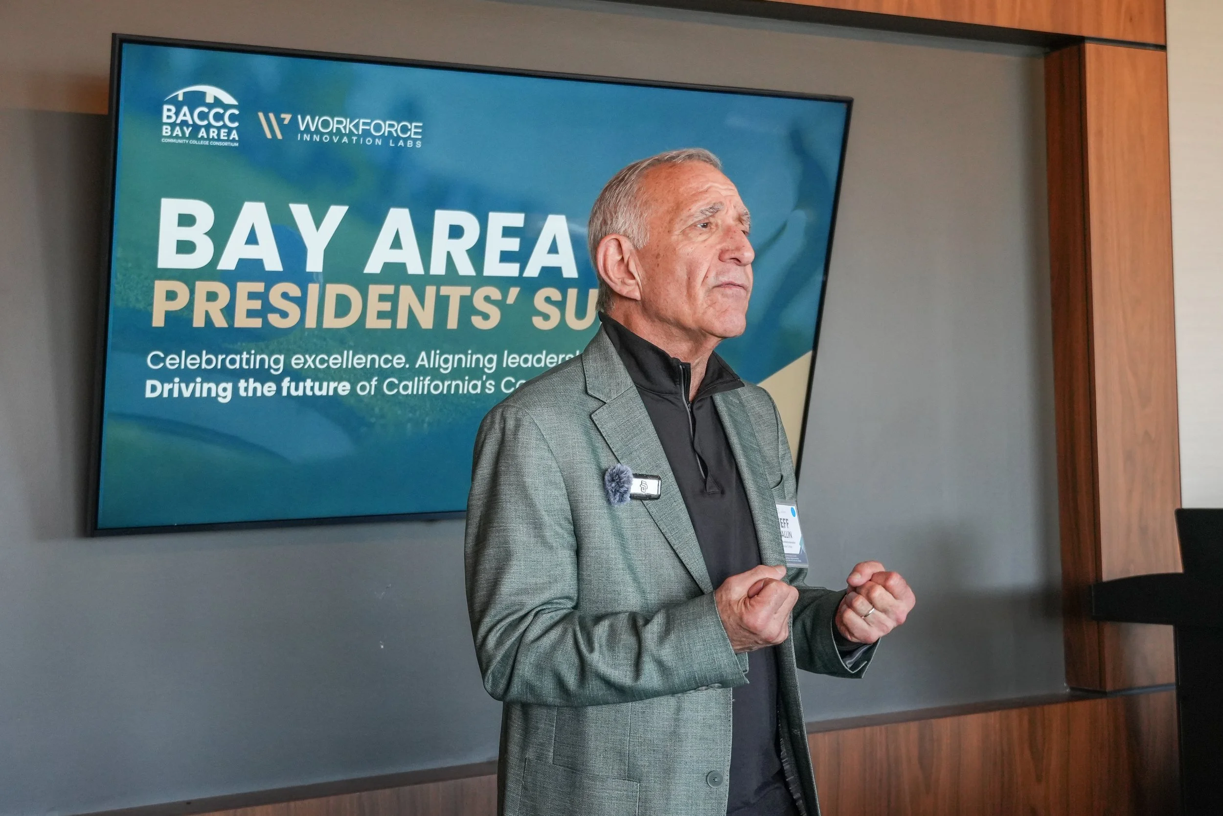 A man in a gray blazer and black shirt is speaking at a conference, standing in front of a large digital screen displaying a BAY AREA Presidents' Summit banner. The banner includes logos for BACCC and Workforce Innovation Labs, and the slogan 'Celebr