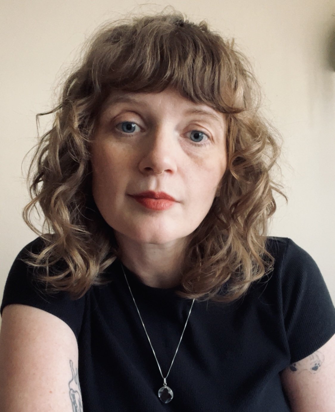 A woman with curly light brown hair, blue eyes, red lipstick, wearing a black shirt and a silver necklace with a pendant, looking at the camera.