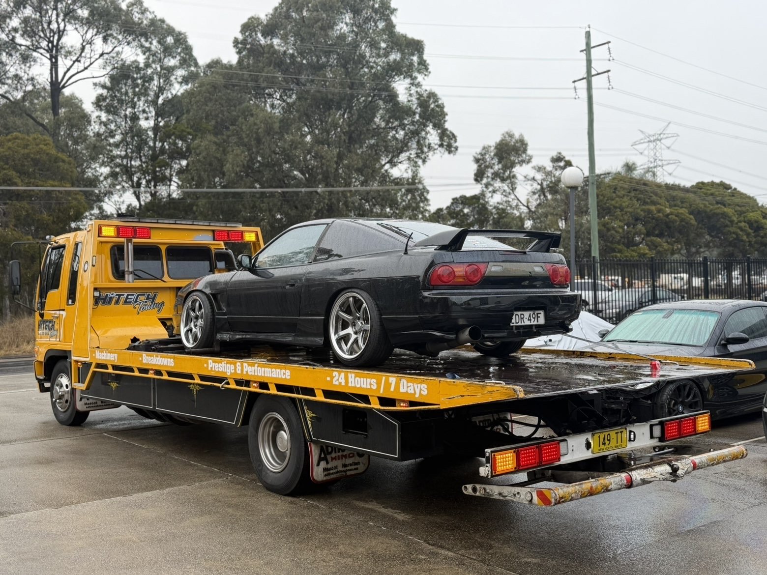 Black sports car on yellow flatbed tow truck in a parking lot, rainy weather.