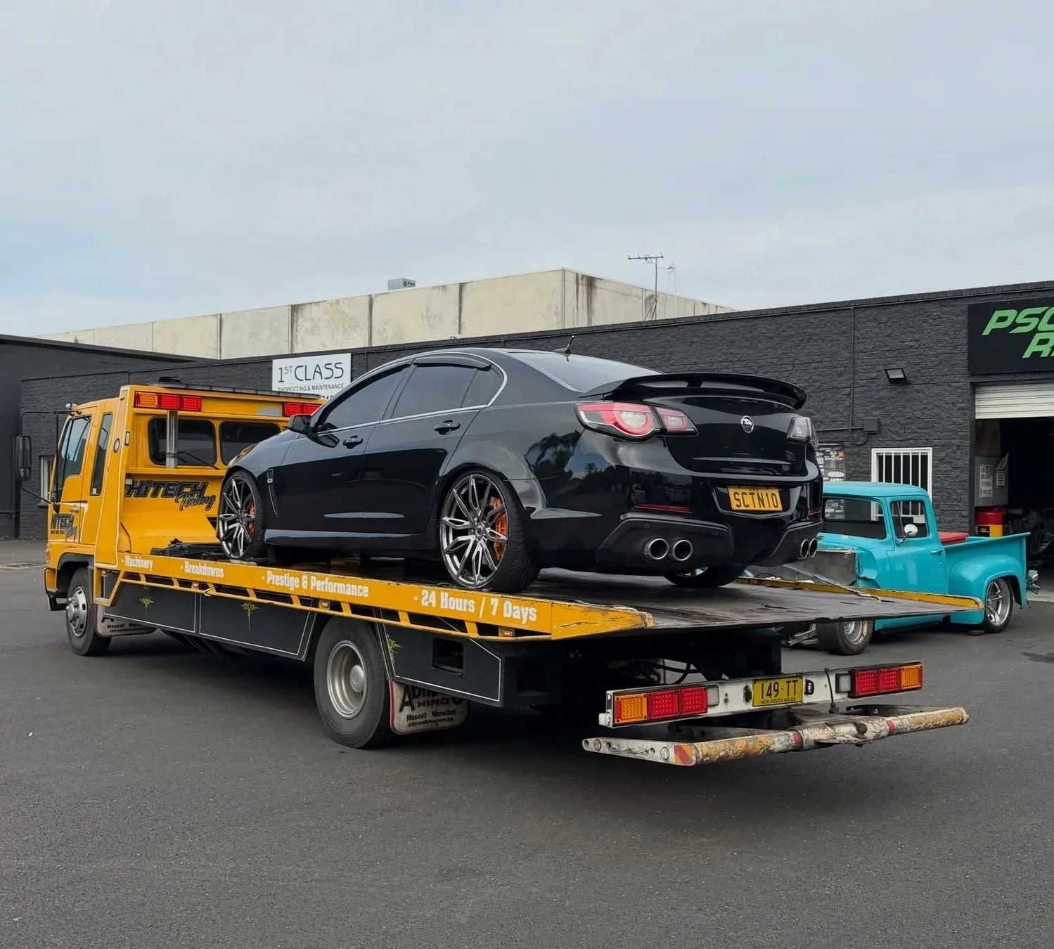Black sports car with custom wheels loaded on a yellow flatbed tow truck in a parking lot, with a blue vintage pickup truck nearby, and black buildings in the background.