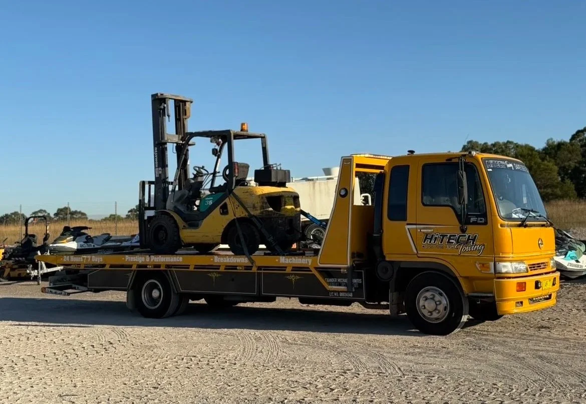 A yellow tow truck carrying a forklift and a jet ski on a flatbed trailer, parked outdoors on a dirt surface with a clear blue sky.