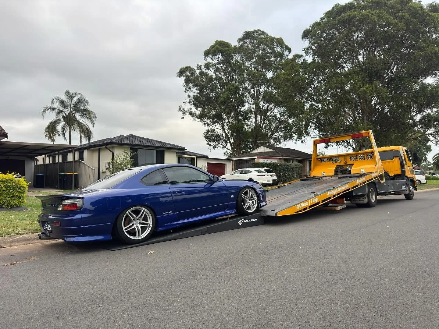 A blue sports car being loaded onto a yellow tow truck on a residential street with houses, trees, and parked cars.