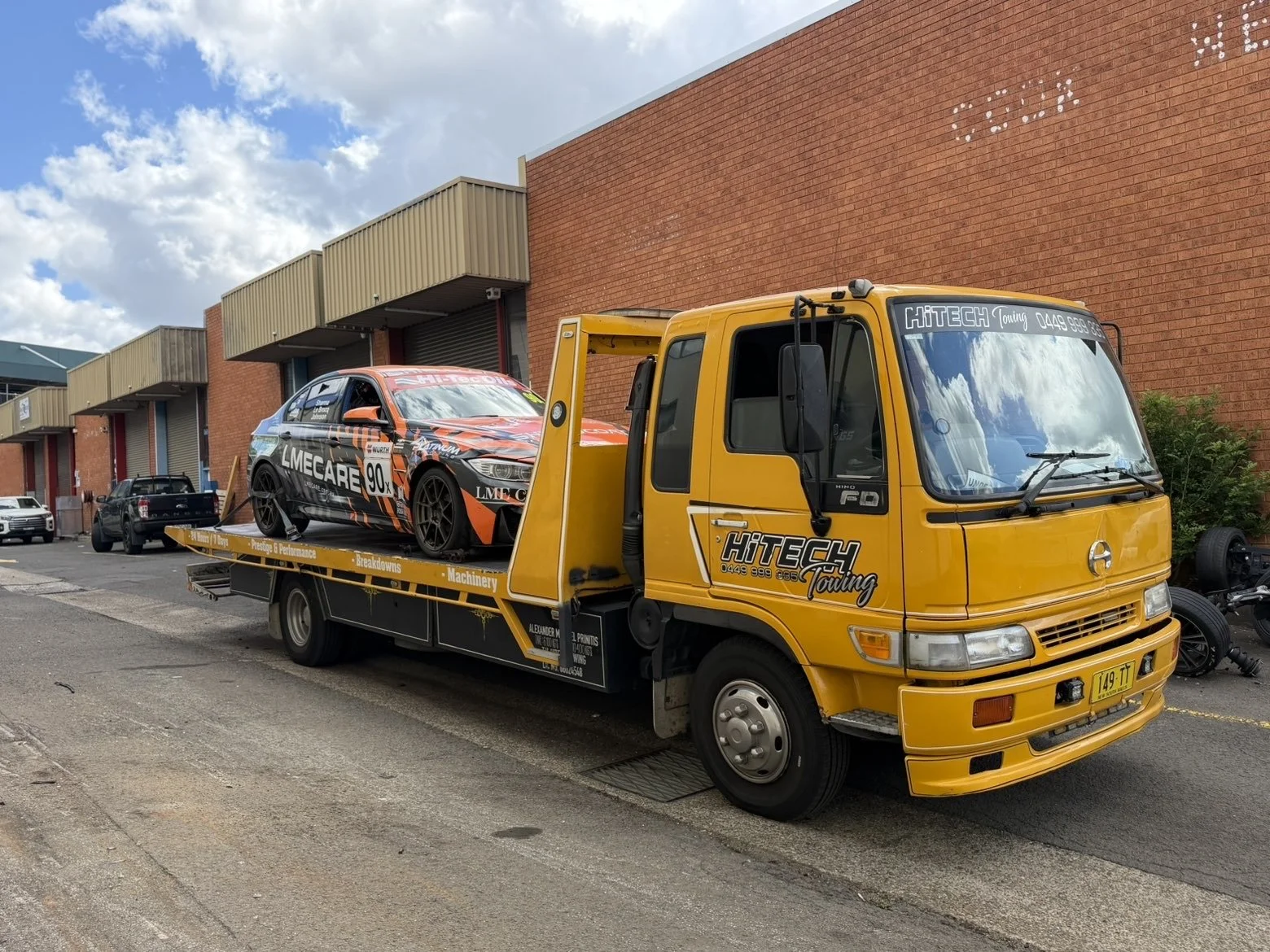 A yellow tow truck from Hi-Tech Towing is parked on the side of the street, carrying a racing car with a black, orange, and gray livery.