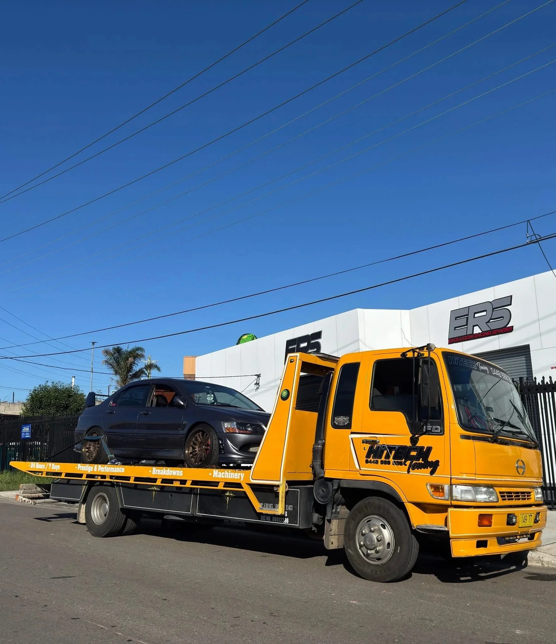 A yellow flatbed tow truck transporting a black car. The truck has branding for Hittech Touring and various service features listed on the side. The background shows a commercial building with signages, power lines in the sky, and sunny weather.