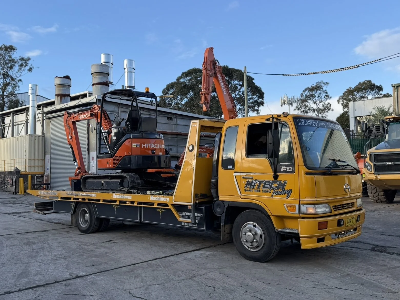 A yellow towing truck with branding 'Hitech Towing' on the side is transporting a small orange excavator. The scene is outdoors on a paved surface with industrial buildings and trees in the background.