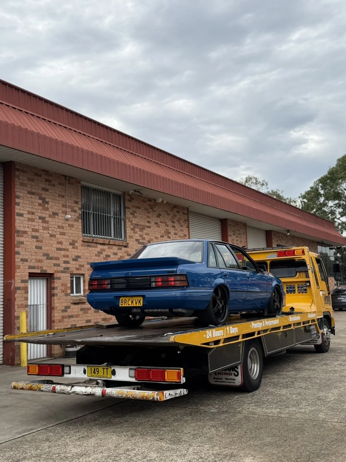 A blue car on a yellow tow truck parked on a concrete driveway in front of a brick building with a red metal roof.
