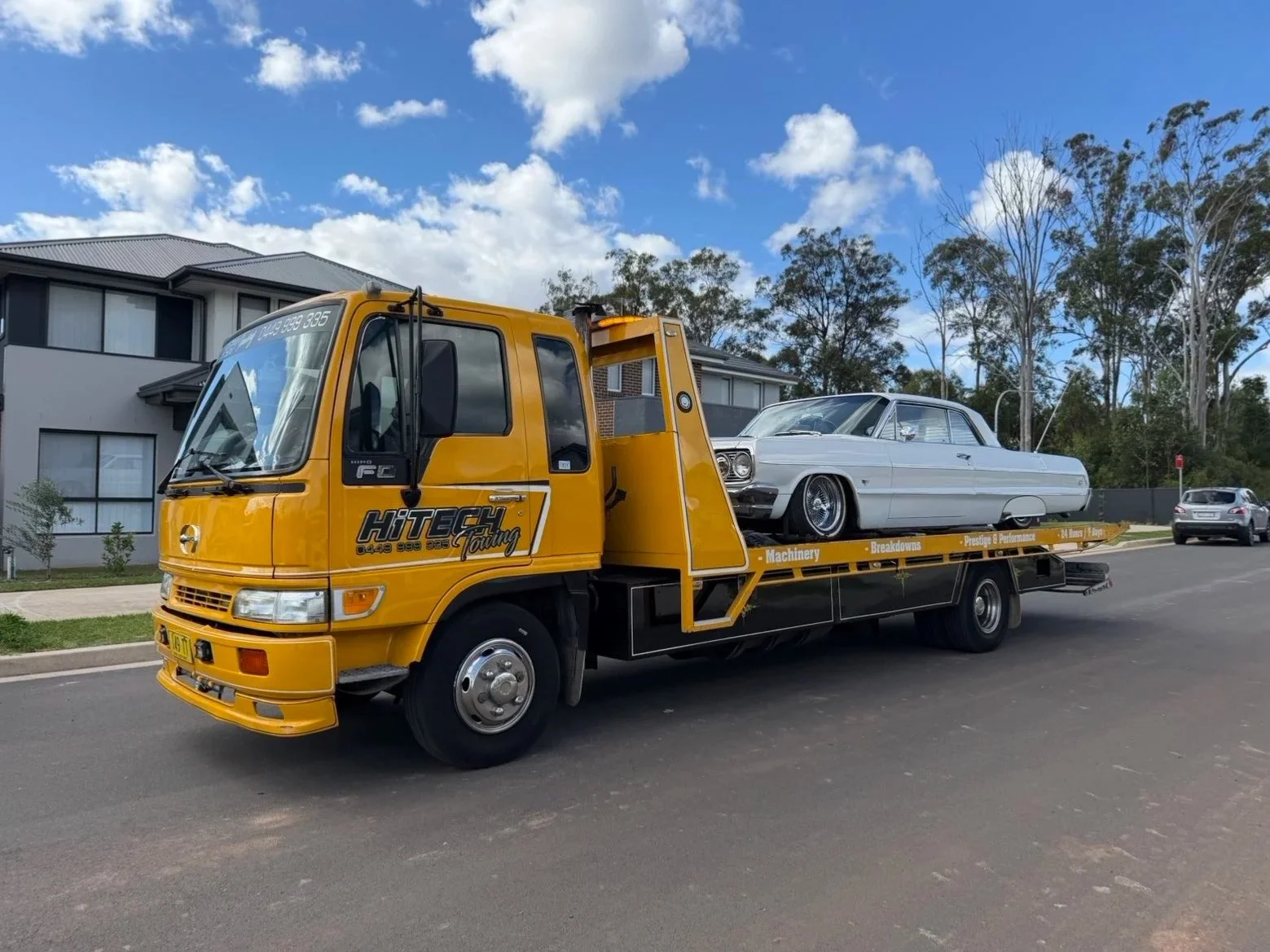 A yellow tow truck carrying a classic white car on its flatbed, parked on a residential street with modern houses and trees under a partly cloudy sky.