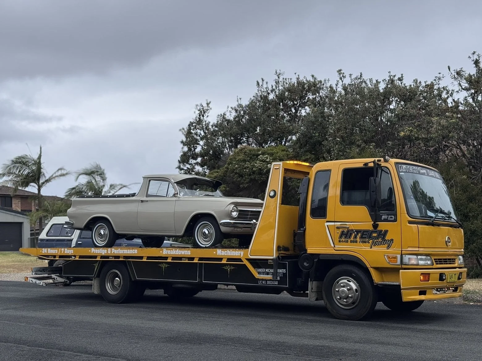 A yellow tow truck carrying a vintage beige pickup truck on its flatbed. The truck has the words 'HI TECH Towing' and 'Prestige & Performance - Breakdowns - Machinery' written on the side, parked on a street with trees and houses in the background under a cloudy sky.