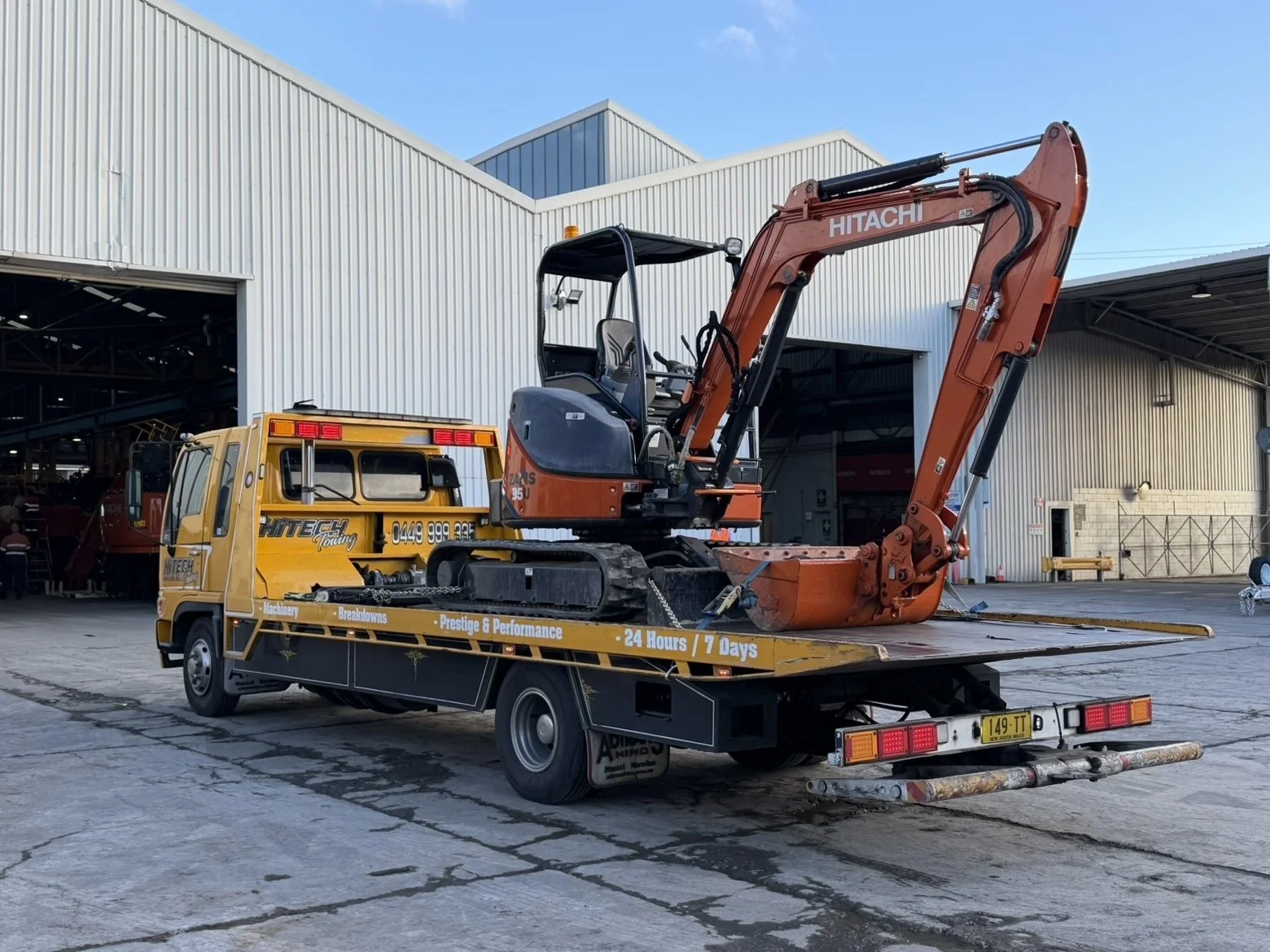 A small orange excavator on a yellow flatbed truck in an industrial yard outside a large warehouse. The flatbed has company branding and contact details, with a black and gray excavator with a bucket attachment.