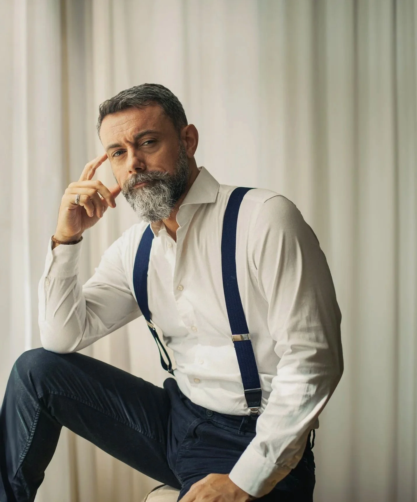 A middle-aged man with a gray beard, wearing a white dress shirt, dark pants, blue suspenders, and a wristwatch, sitting with one leg up and touching his temple with his hand, looking at the camera with an inquisitive expression in front of a light curtain background.