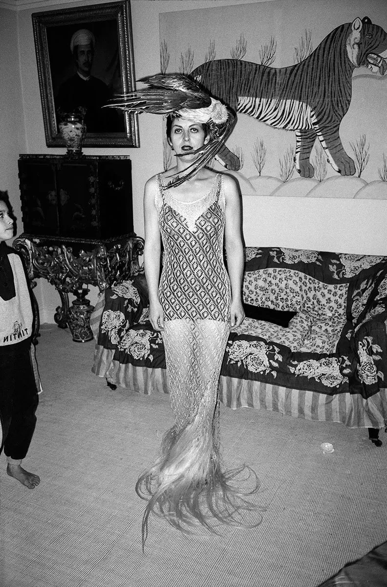 A woman with a mermaid tail and a bird on her head standing in a living room with a tiger mural on the wall, a sofa with patterned cushions, a framed portrait on the wall, and a boy standing nearby.