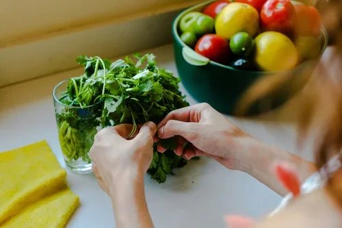 Person washing fresh cilantro in a glass bowl on a kitchen counter with a bowl of colorful apples in the background.