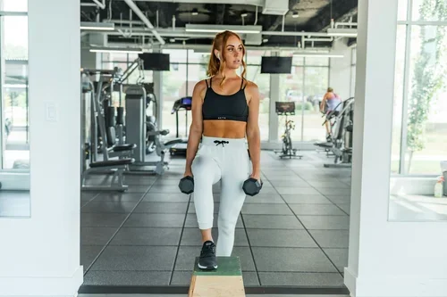 A woman in workout clothing holding dumbbells in a gym, standing on a wooden balance board.