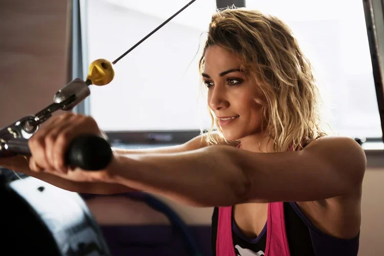 A young woman with curly blonde hair, wearing a sports top and pink workout vest, is exercising with a rowing machine in a fitness center.