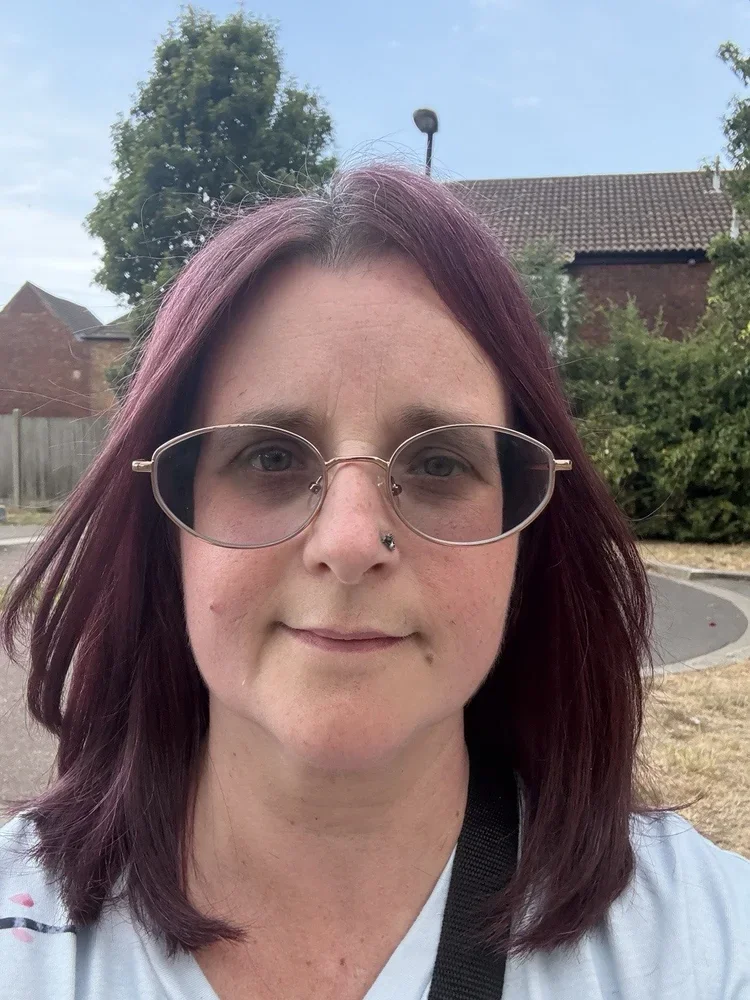 A woman with purple hair and glasses taking a selfie outdoors, with trees, a brick house, and a cloudy sky in the background.