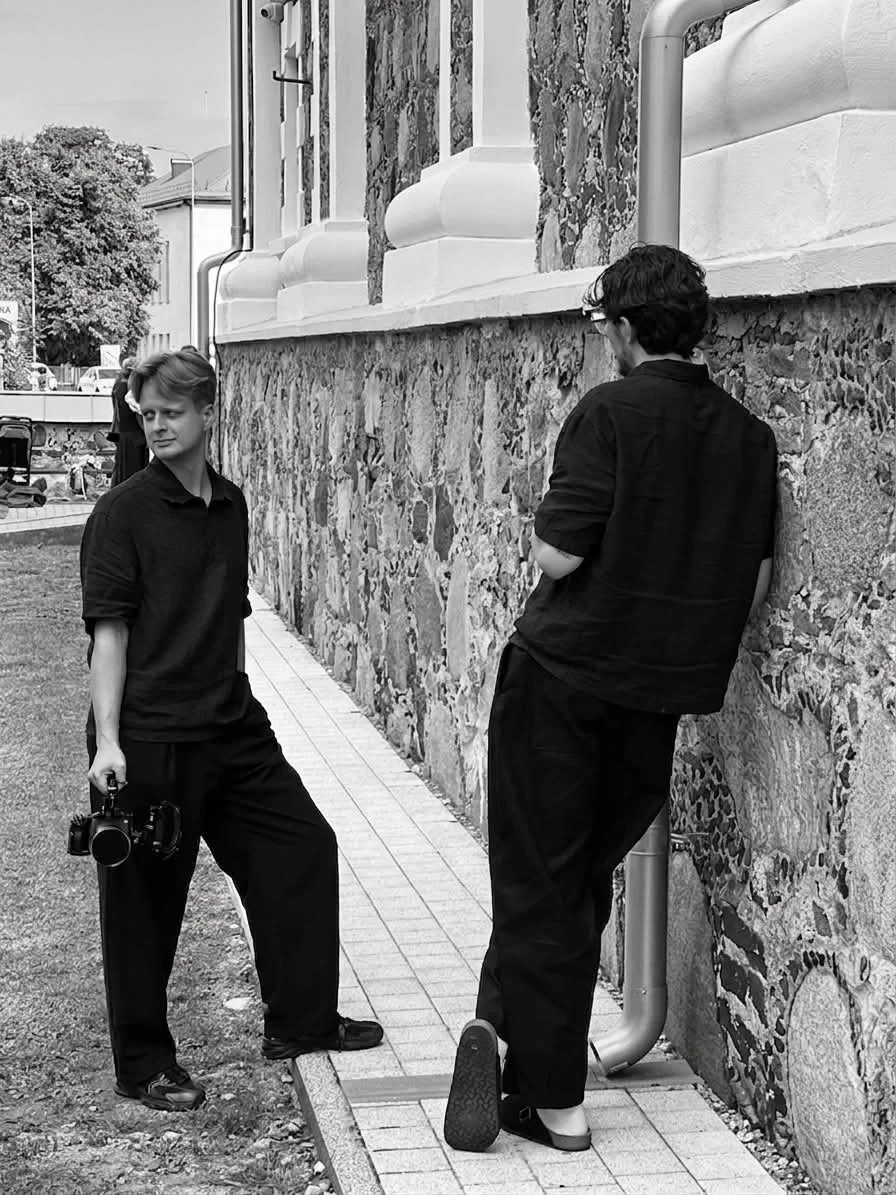 Two young men in black clothing, one holding a camera, stand outdoors near a stone building with a patterned wall. One leans against the wall with arms crossed, the other stands with one foot on a low stone edge, looking away. Filming weddings.