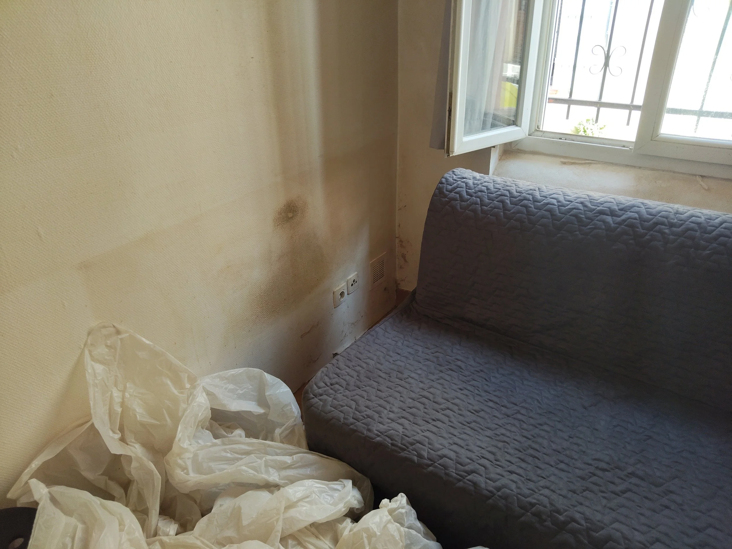 Mur sale avec taches d'humidité, un fauteuil en tissu gris, une fenêtre ouverte laissant entrer de la lumière, et un tas de plastique blanc sur le sol.