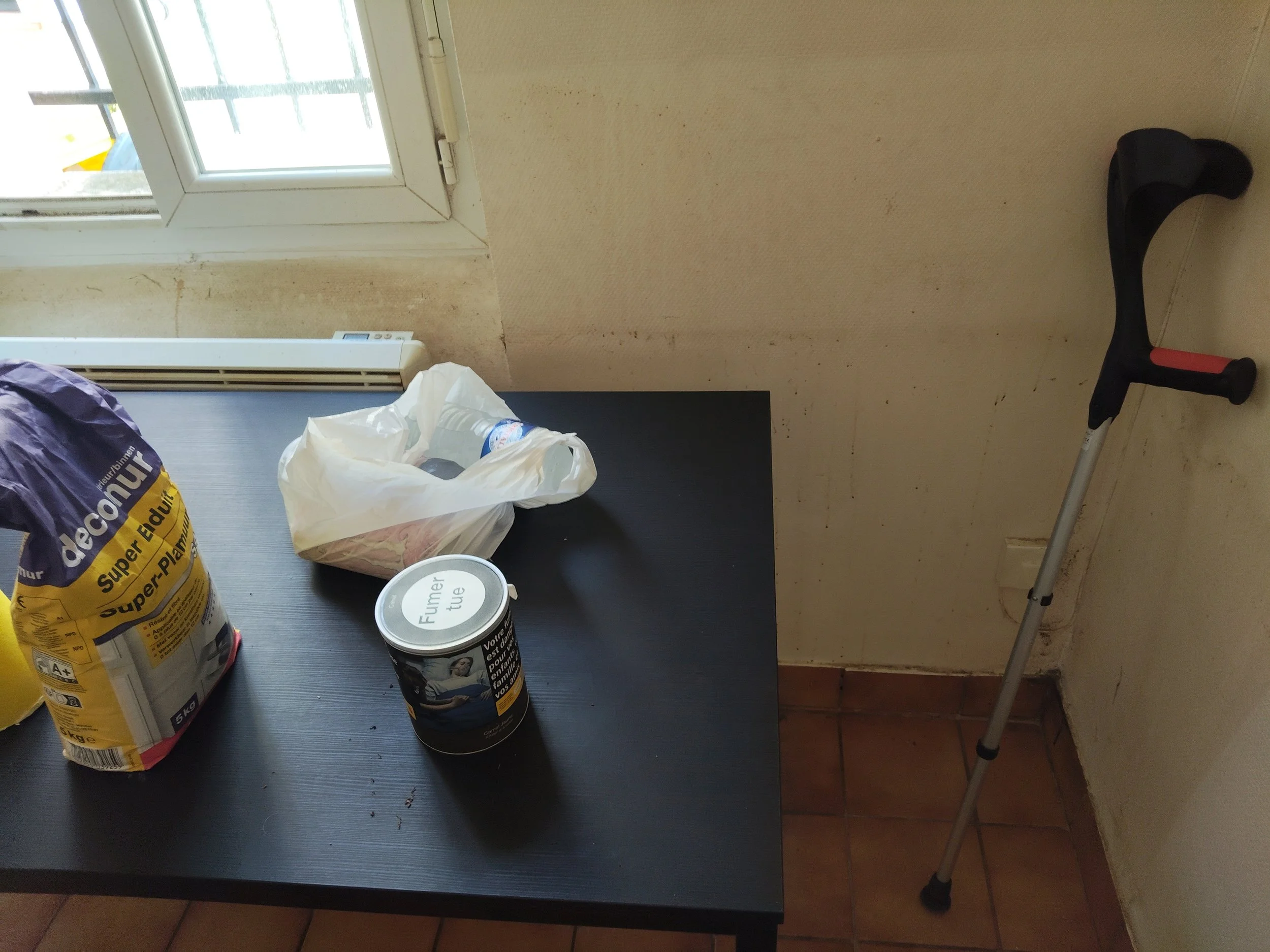 Une table formée de quatre sacs de nourriture, deux canettes, un sac en plastique blanc, une marchette et un radiateur. La pièce a un mur beige avec des taches et une fenêtre qui laisse entrer la lumière.