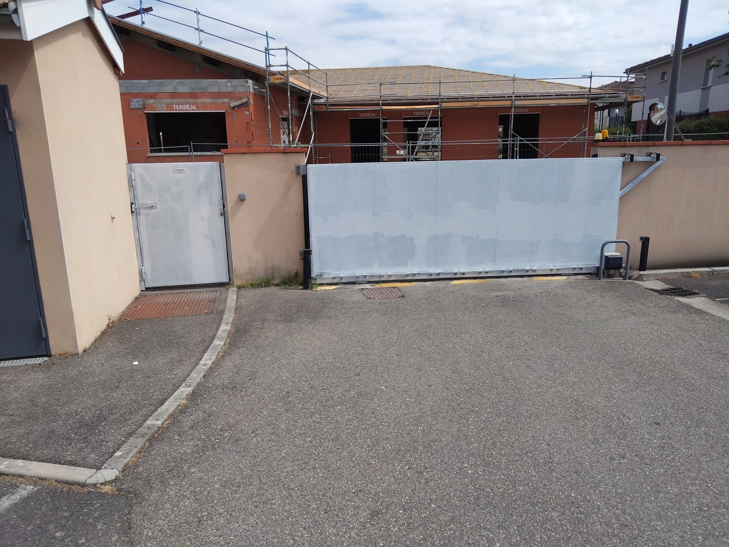 Construction d'une maison avec échafaudages, murs en briques et toiture en cours de finition. Un portail métallique blanc ferme l'entrée, et un mur avec un portail et une porte en métal est présent au premier plan.