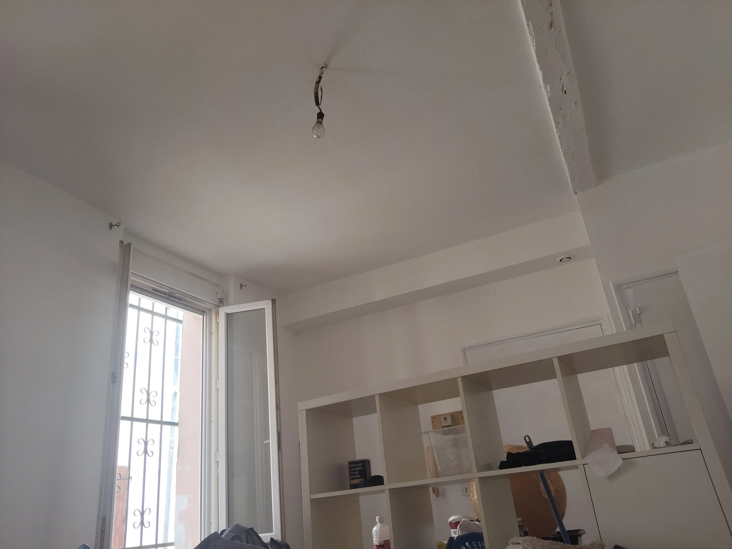 Intérieur d'une pièce en rénovation avec plafond en cours de travaux, lumière suspendue non installée, meubles en cours de montage, grande fenêtre laissant entrer la lumière naturelle.
